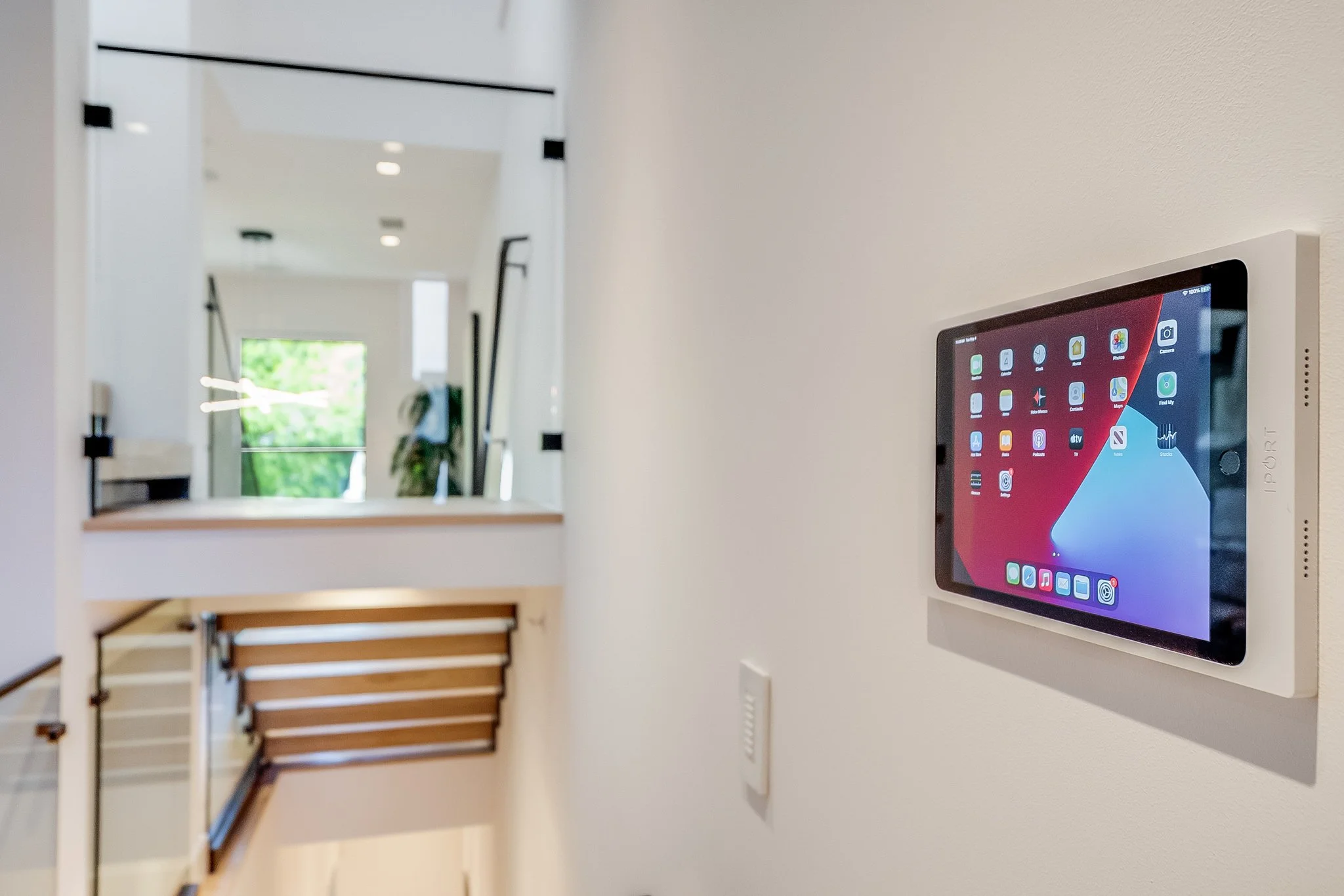 Interior of a modern home with a wall-mounted iPad displaying a home screen, next to a staircase with wooden steps and a white wall.
