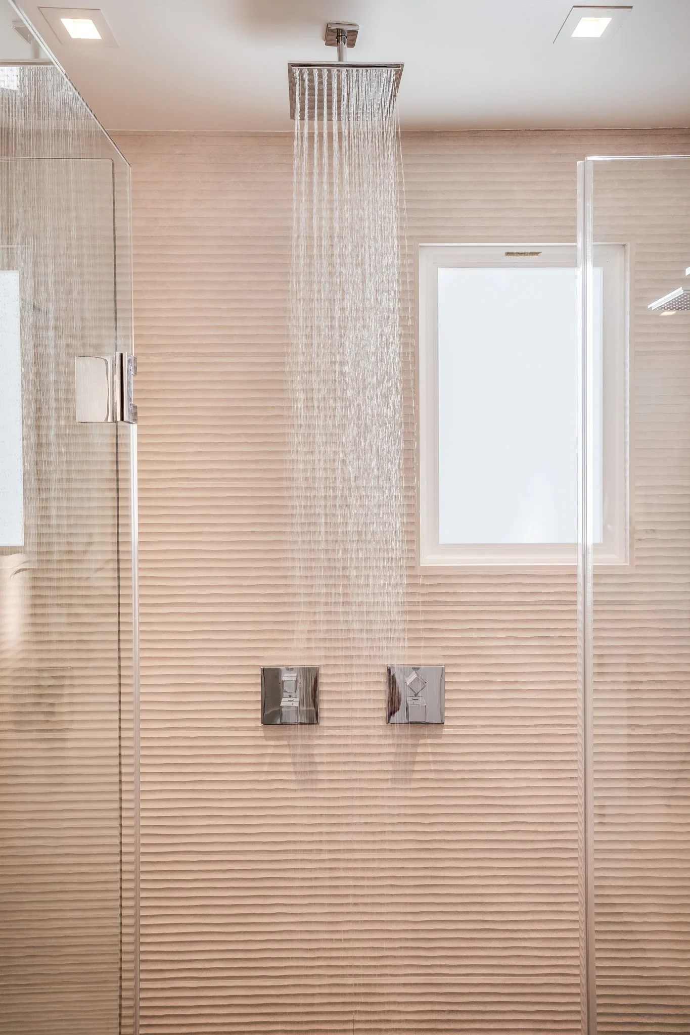 A modern shower with a rainfall showerhead, beige textured wall tiles, a window, and glass shower door.