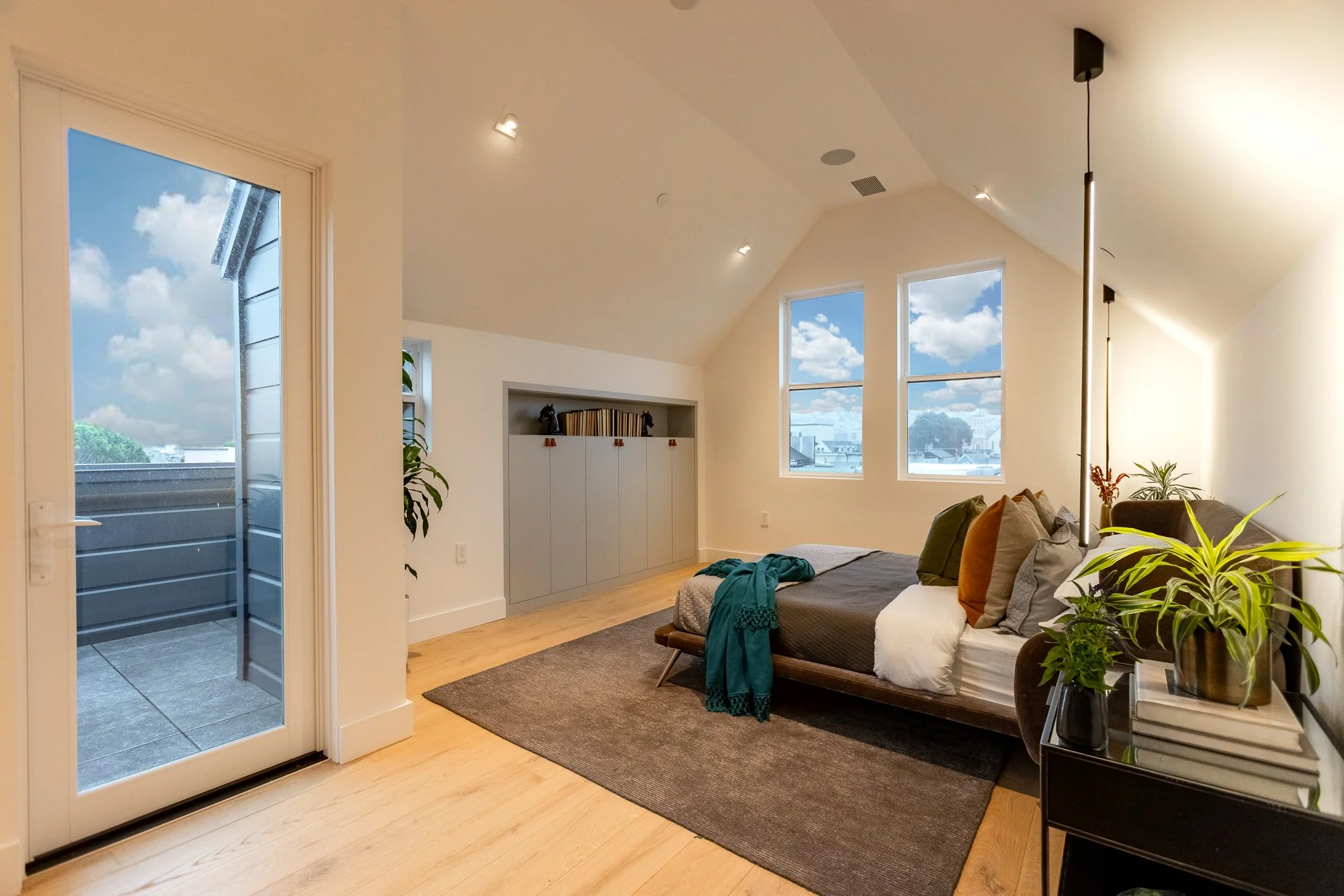 Modern bedroom with large windows showing a blue sky with clouds, a door leading to a balcony, a bed with multiple pillows and a blanket, a small black table with plants and books, and a built-in cabinet with decorations.