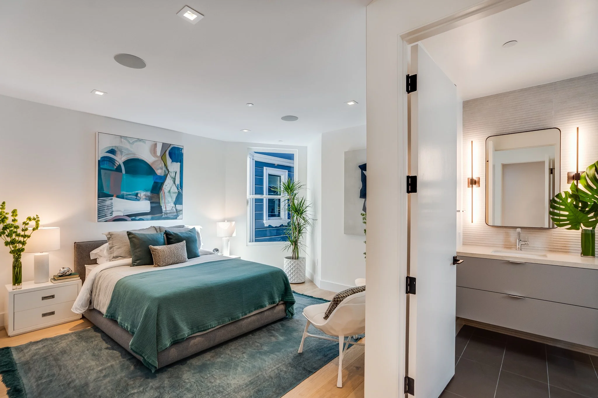 Modern bedroom with bed, abstract art on the wall, green bedding, nightstand with lamp, and a window with blue exterior.