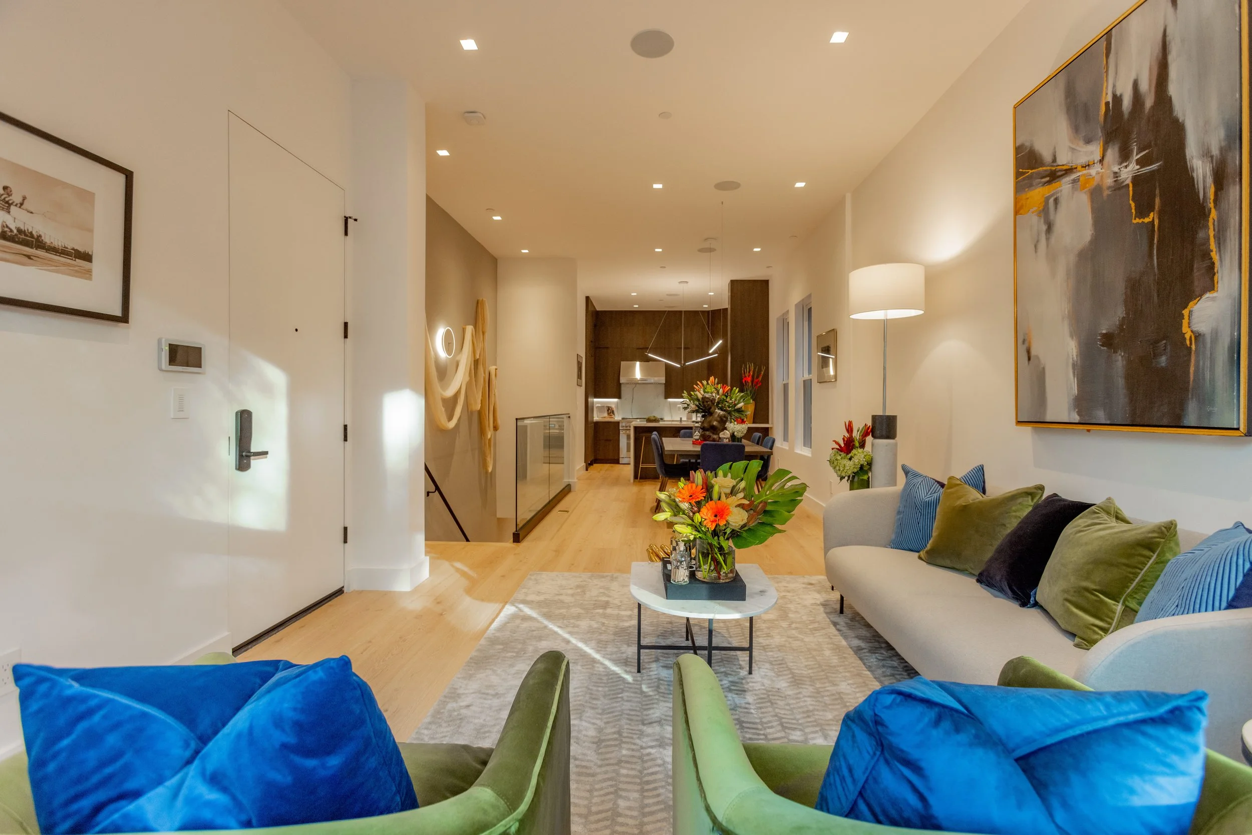Living room with a beige sofa, green and blue chairs, colorful pillows, and a coffee table with flower arrangements. Adjacent to a dining area and open kitchen, decorated with modern art and warm lighting.