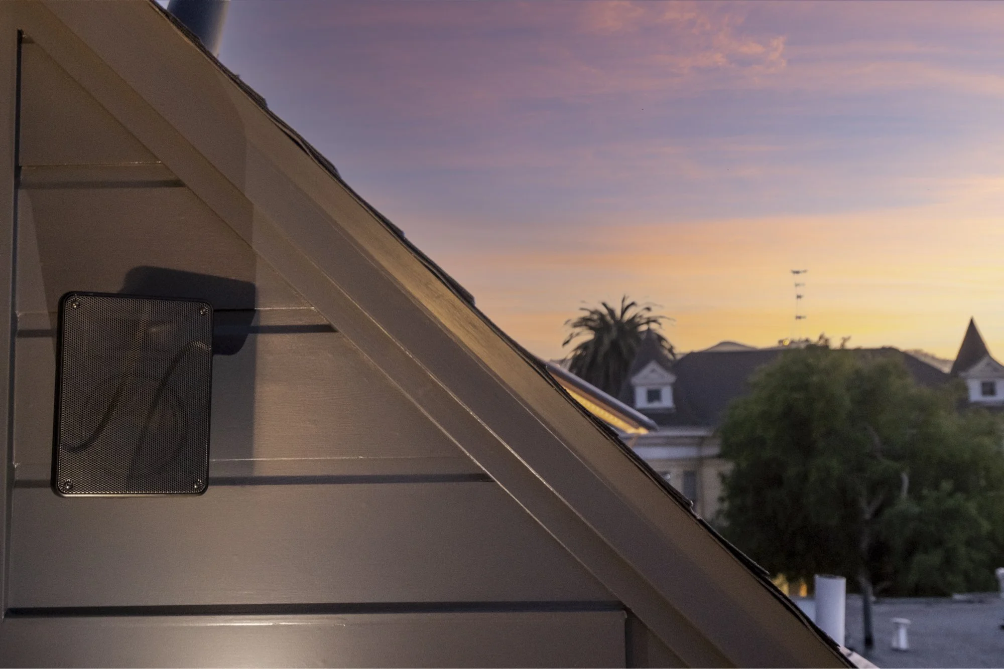 Close-up of the side of a house with a vent, slanted roof, and a sunset sky in the background along with trees and rooftops.