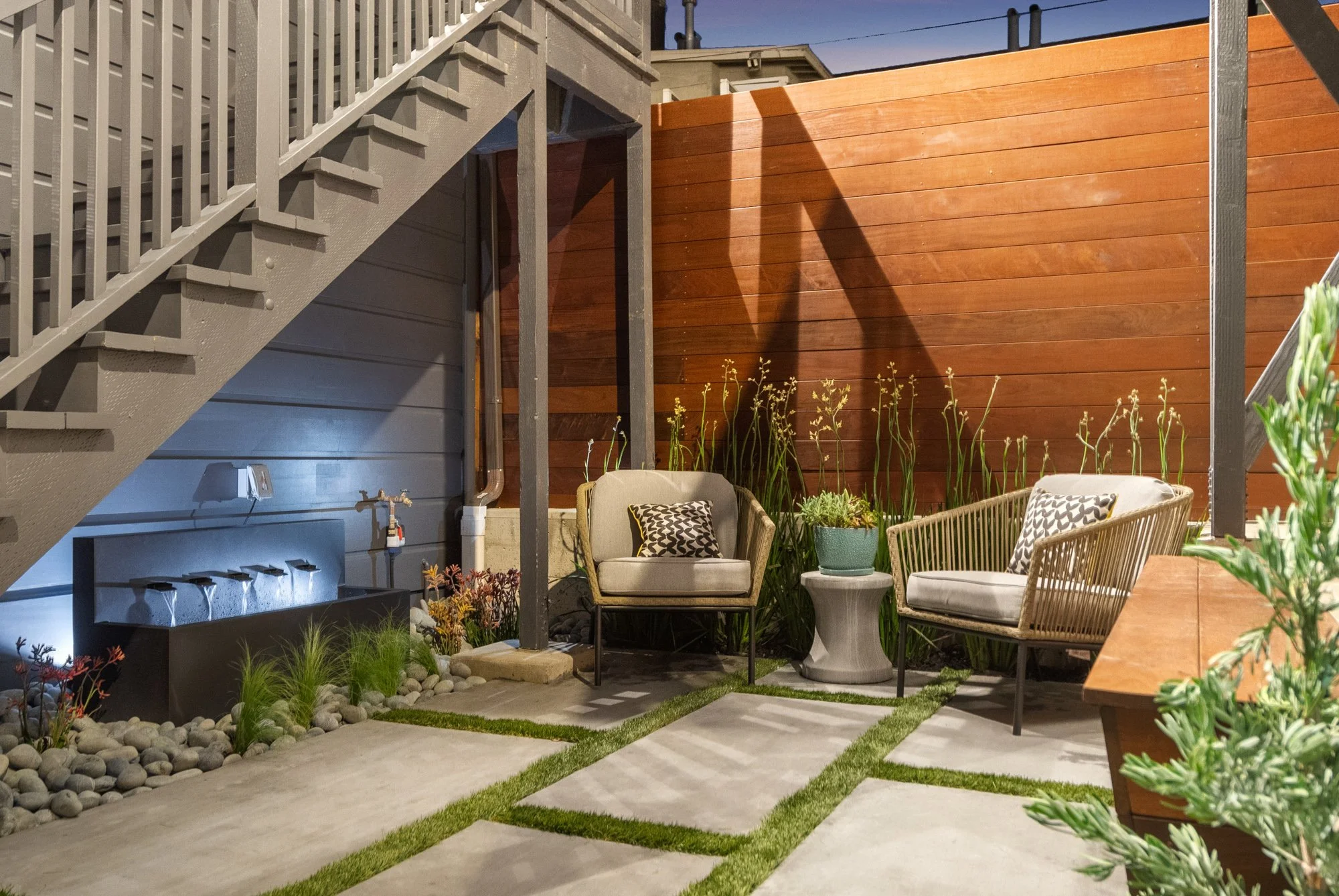 Outdoor patio area with two cushioned chairs, a small side table with a potted plant, a wooden bench, and a water feature. The space is enclosed by a blue wall and a brown wooden fence, with a staircase leading up on the left side of the image.