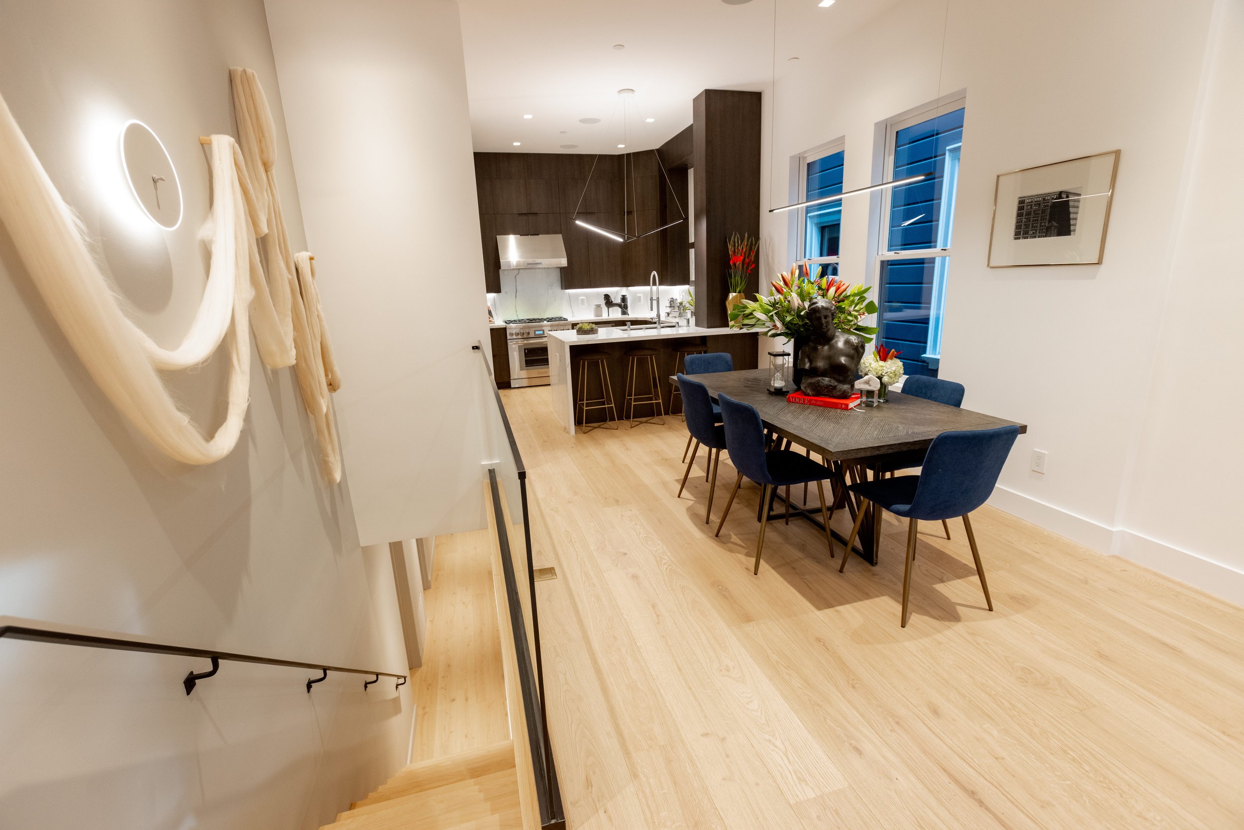 Modern open-concept kitchen and dining area with a dark wood kitchen island, blue dining chairs, a floral centerpiece, and large windows with blue siding outside.