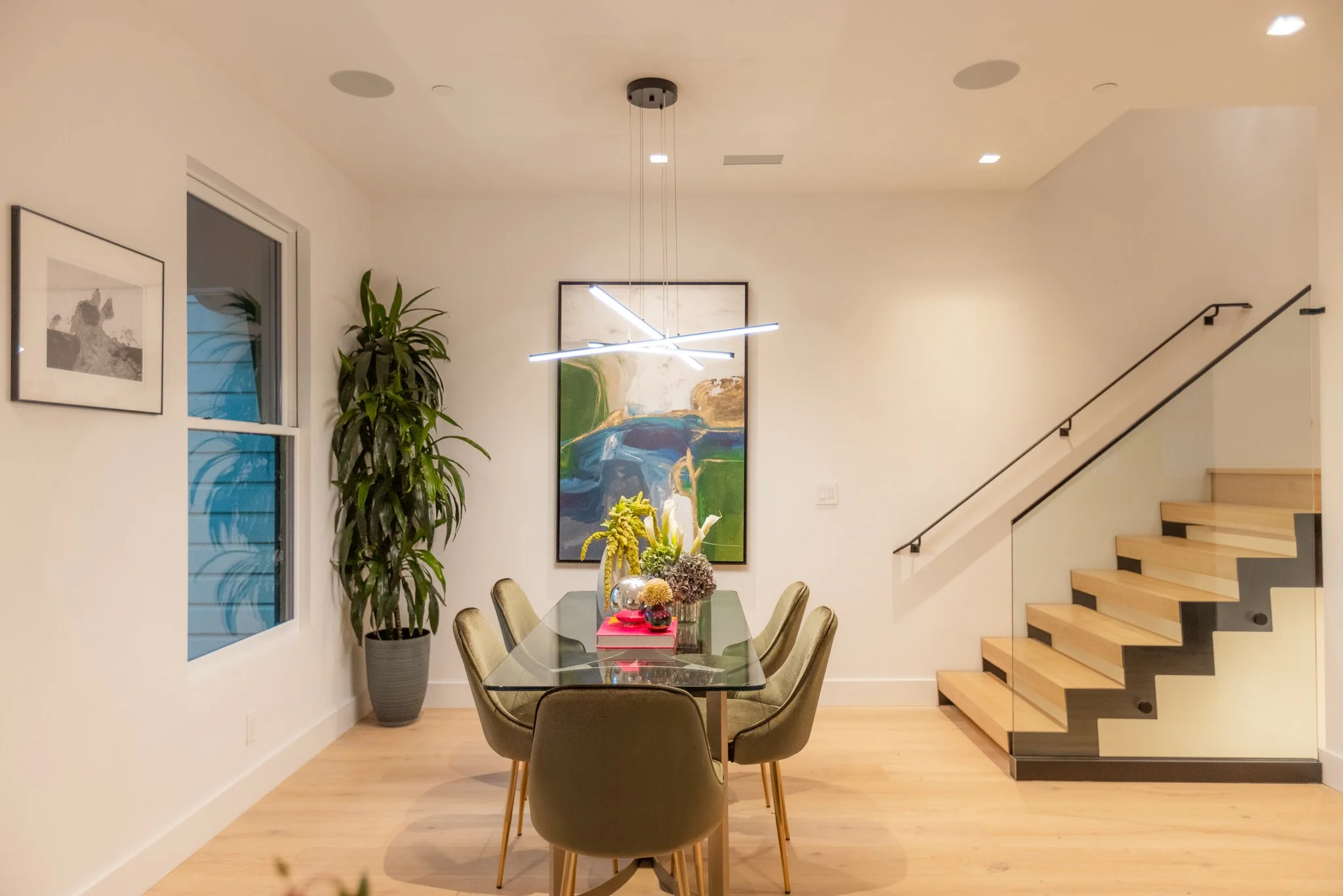 Modern dining room with a rectangular glass table surrounded by six beige chairs with wooden legs. A large potted plant to the left, framed artwork on the wall, and a staircase with wooden steps and black railings on the right. A contemporary chandel