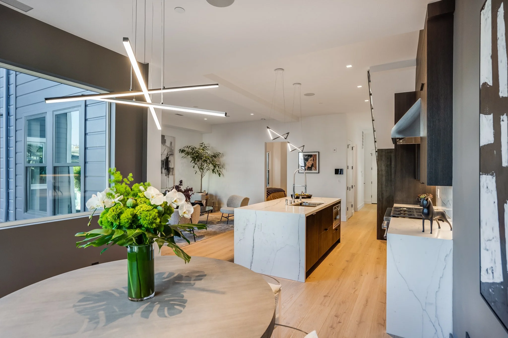 Modern open-concept kitchen with a marble island, contemporary lighting fixtures, and a living area with seating and plants visible in the background.