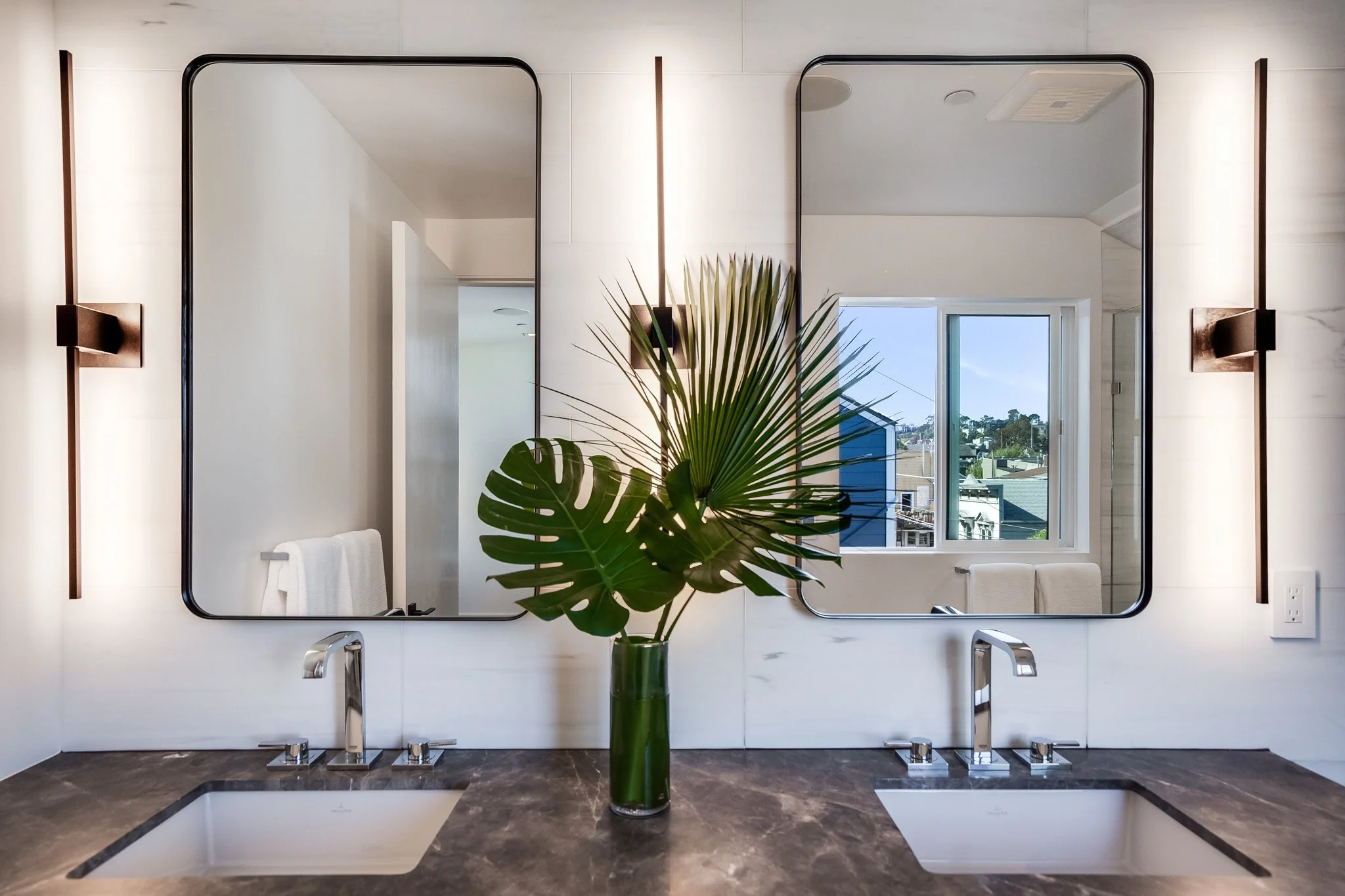 Modern bathroom vanity with dual sinks, a dark marble countertop, two large rectangular mirrors, wall-mounted lights, and a vase with tropical leaves, with a window in the background showing a suburban neighborhood.