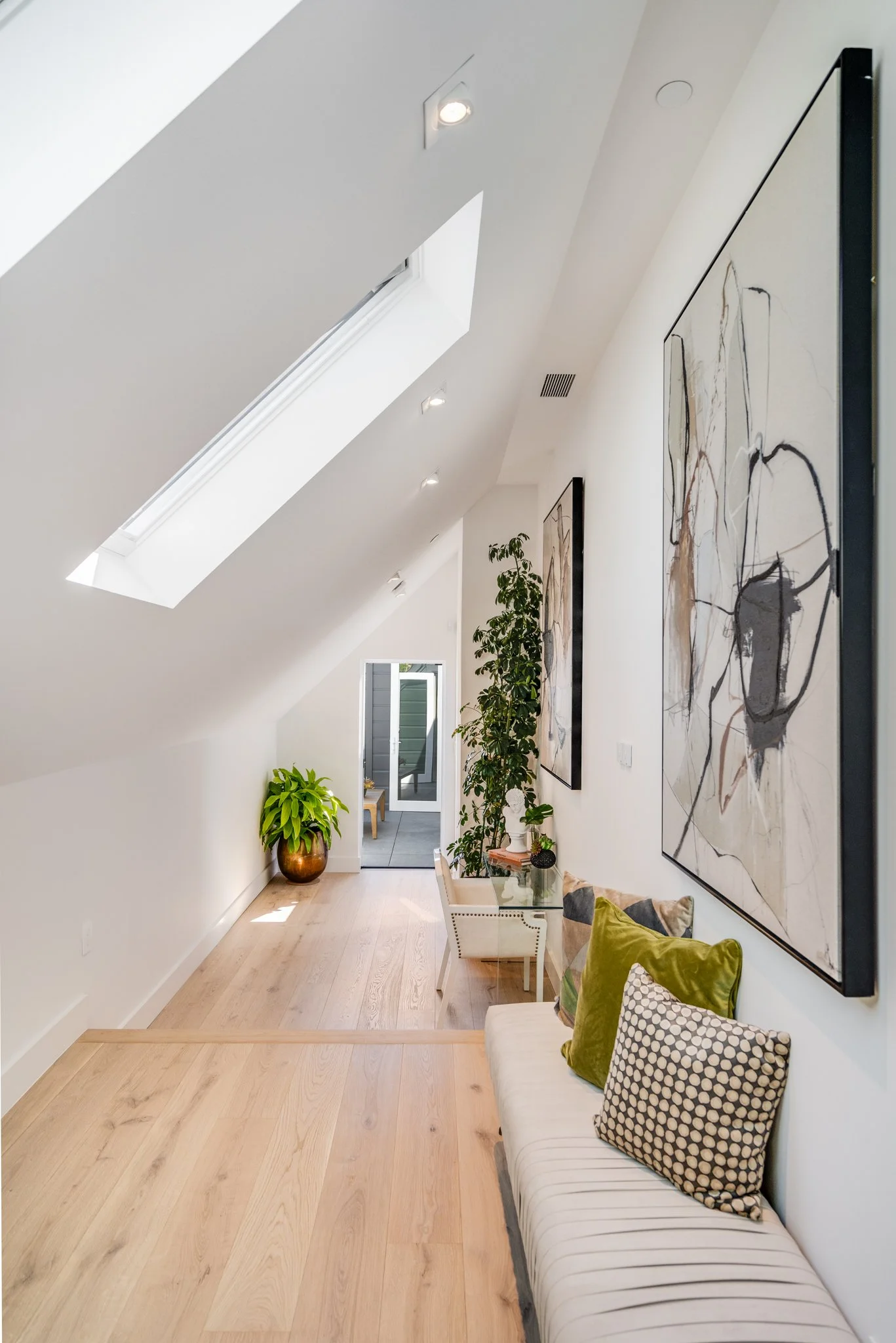 Modern attic room with sloped ceiling, skylight, hardwood flooring, abstract artwork on white walls, potted plants, and a small sitting area with a white couch and decorative pillows.