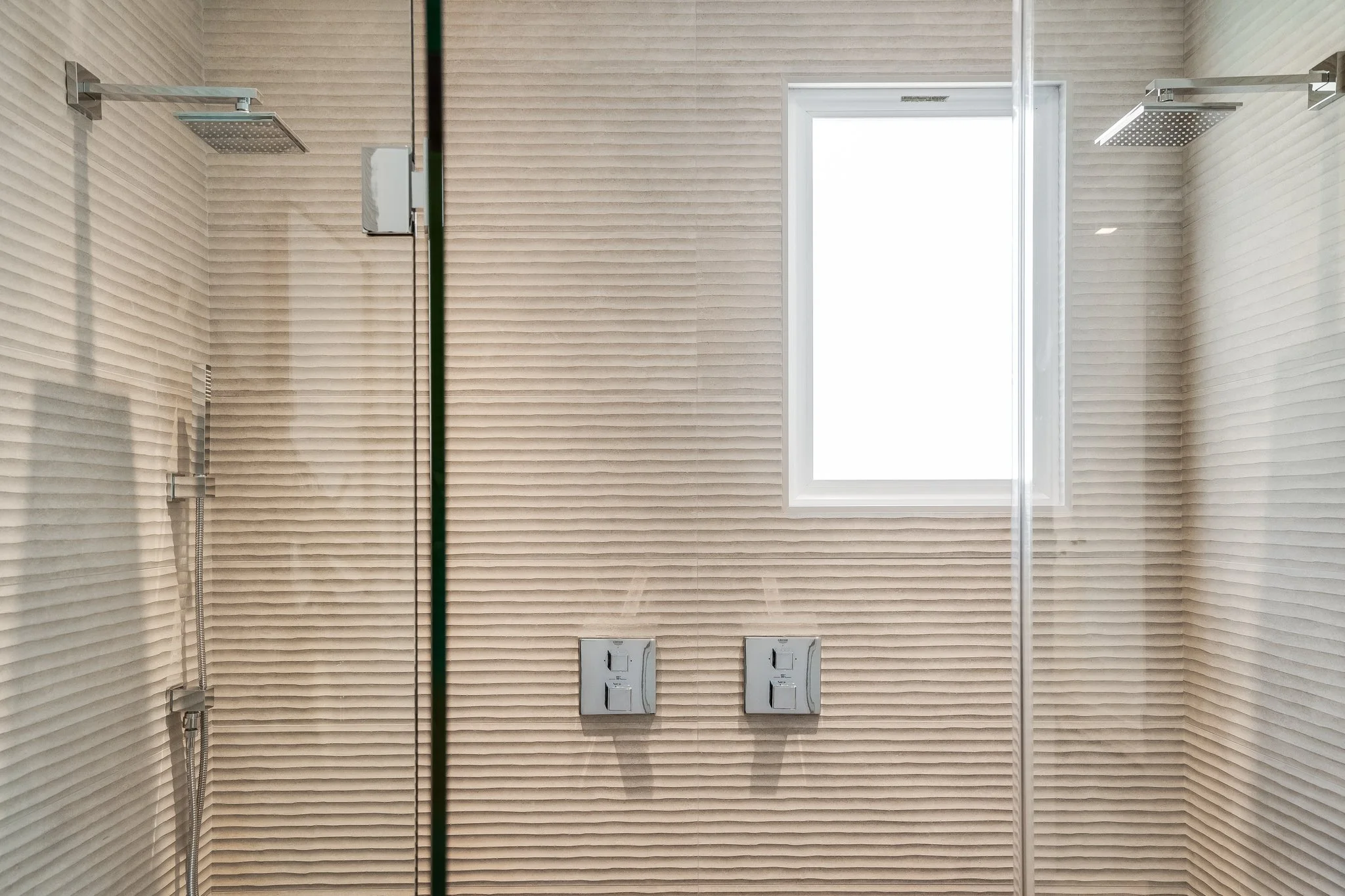 Interior of a modern shower with textured beige tiles, a large window, two showerheads, and two control panels.