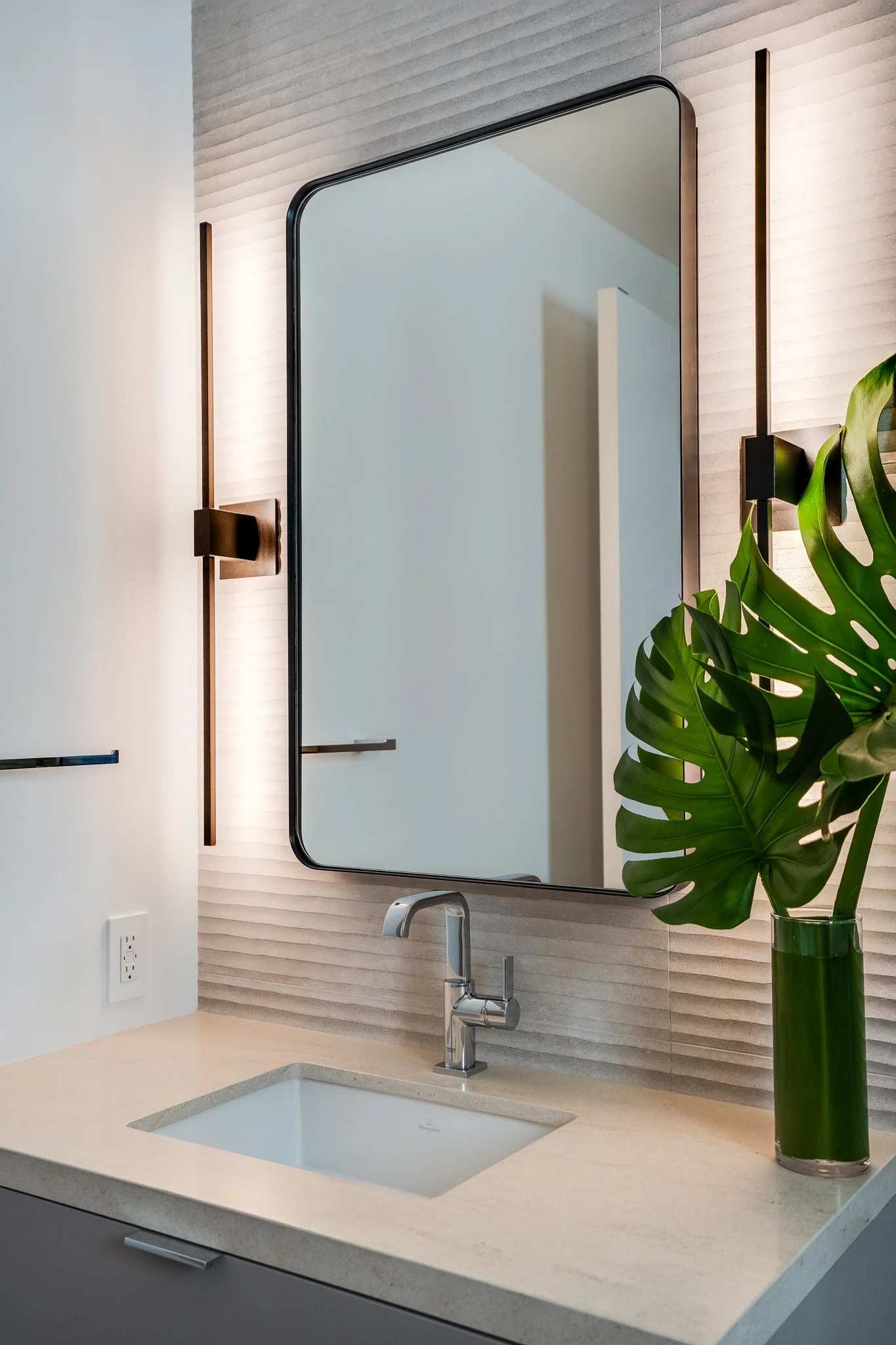 Modern bathroom vanity with a large mirror, a beige countertop, and a sink with a chrome faucet. A green potted plant is on the right side.
