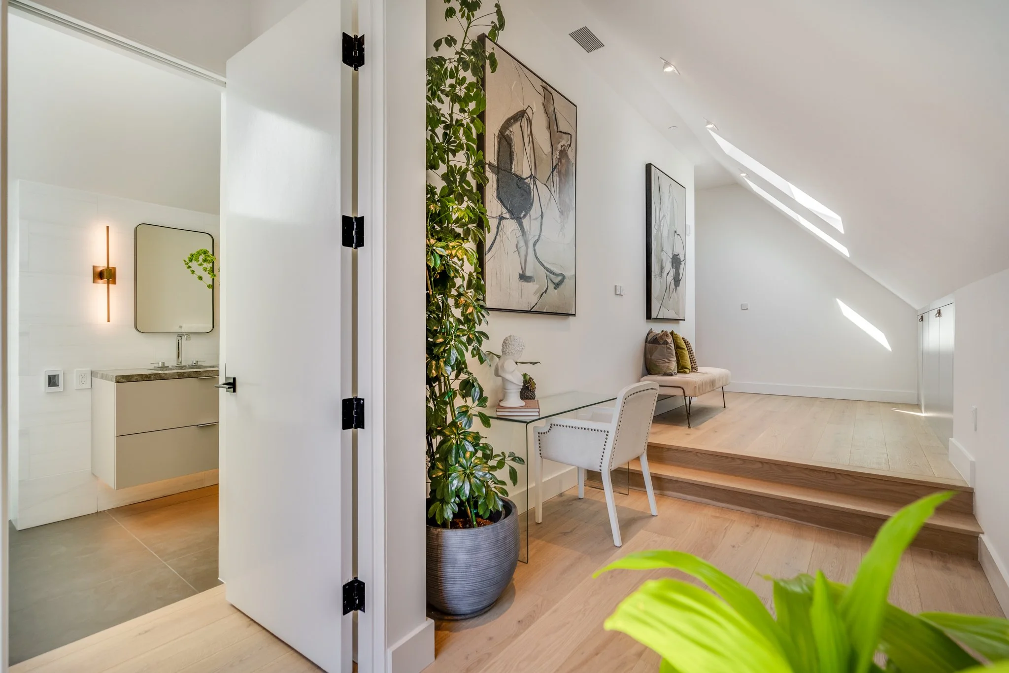 Modern interior living space with a white wall, large abstract artwork, potted plant, and a gray bench with cushions. Skylights on the sloped ceiling provide natural light.