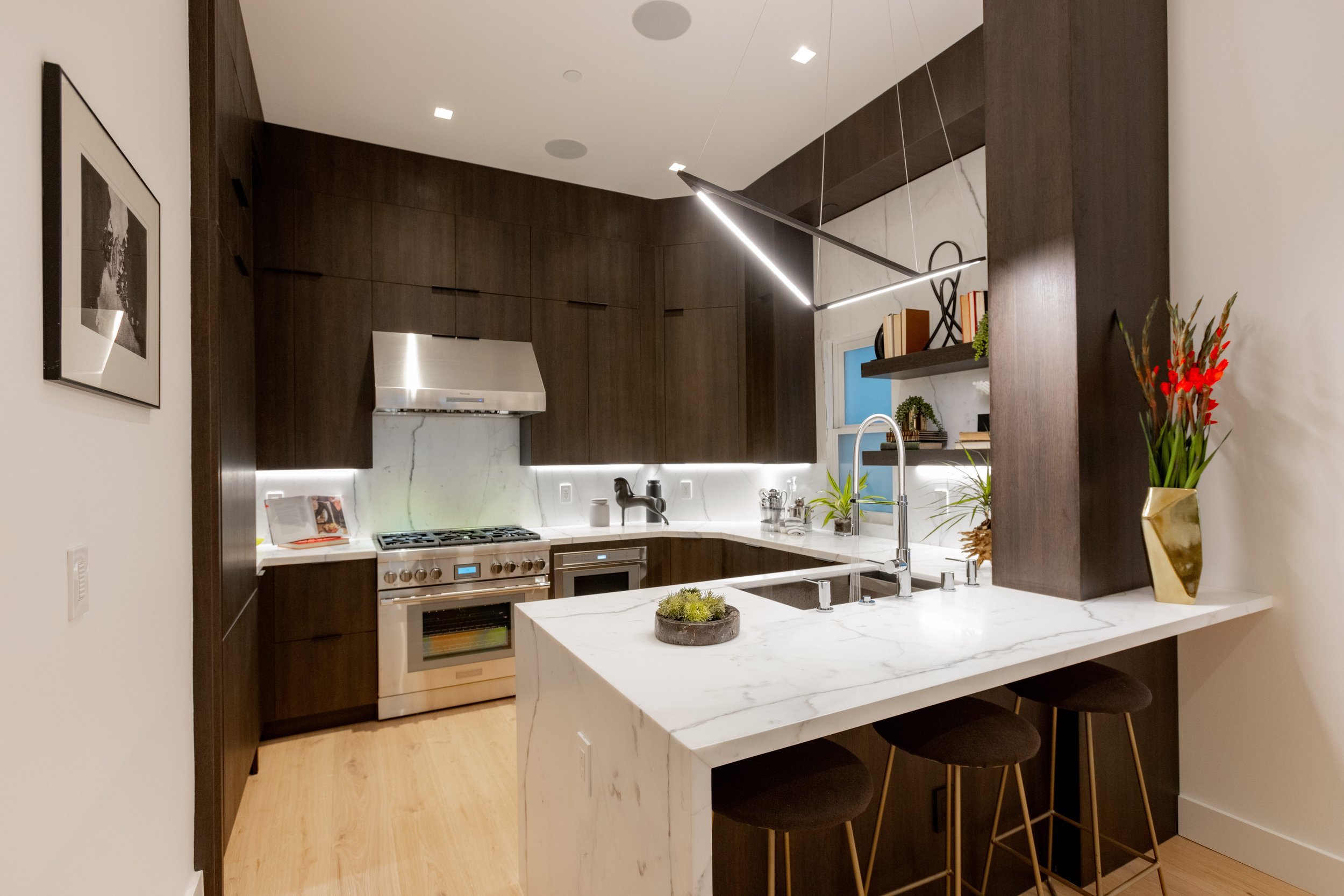 Modern kitchen with dark wood cabinets, white marble countertops, stainless steel appliances, and decorative plants.