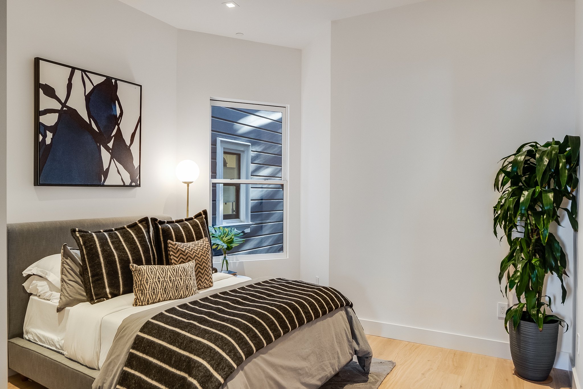 A modern bedroom with a bed that has a gray upholstered headboard, dark striped bedding, and decorative pillows. There is a window with a view of siding outside, a tall potted plant in the corner, a side table with a green plant, and a black-and-whit