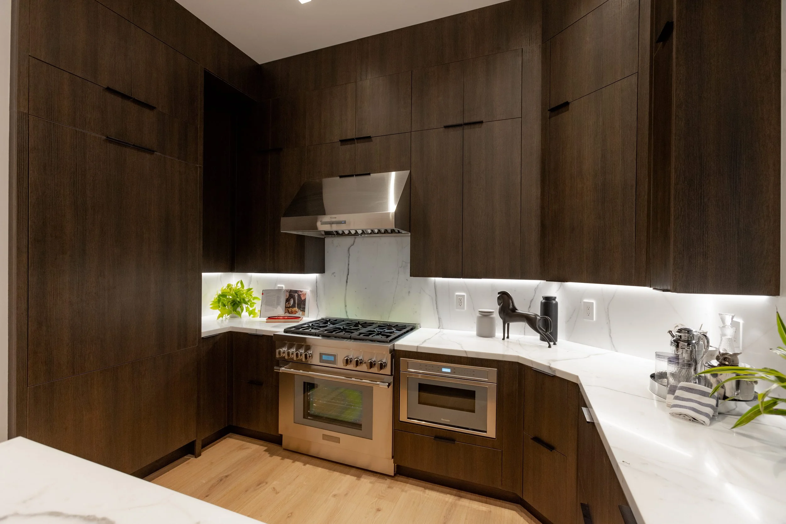 Modern kitchen with dark wood cabinets, white marble countertops, stainless steel appliances, including a stove and oven, and decorative items like a plant and sculptures.