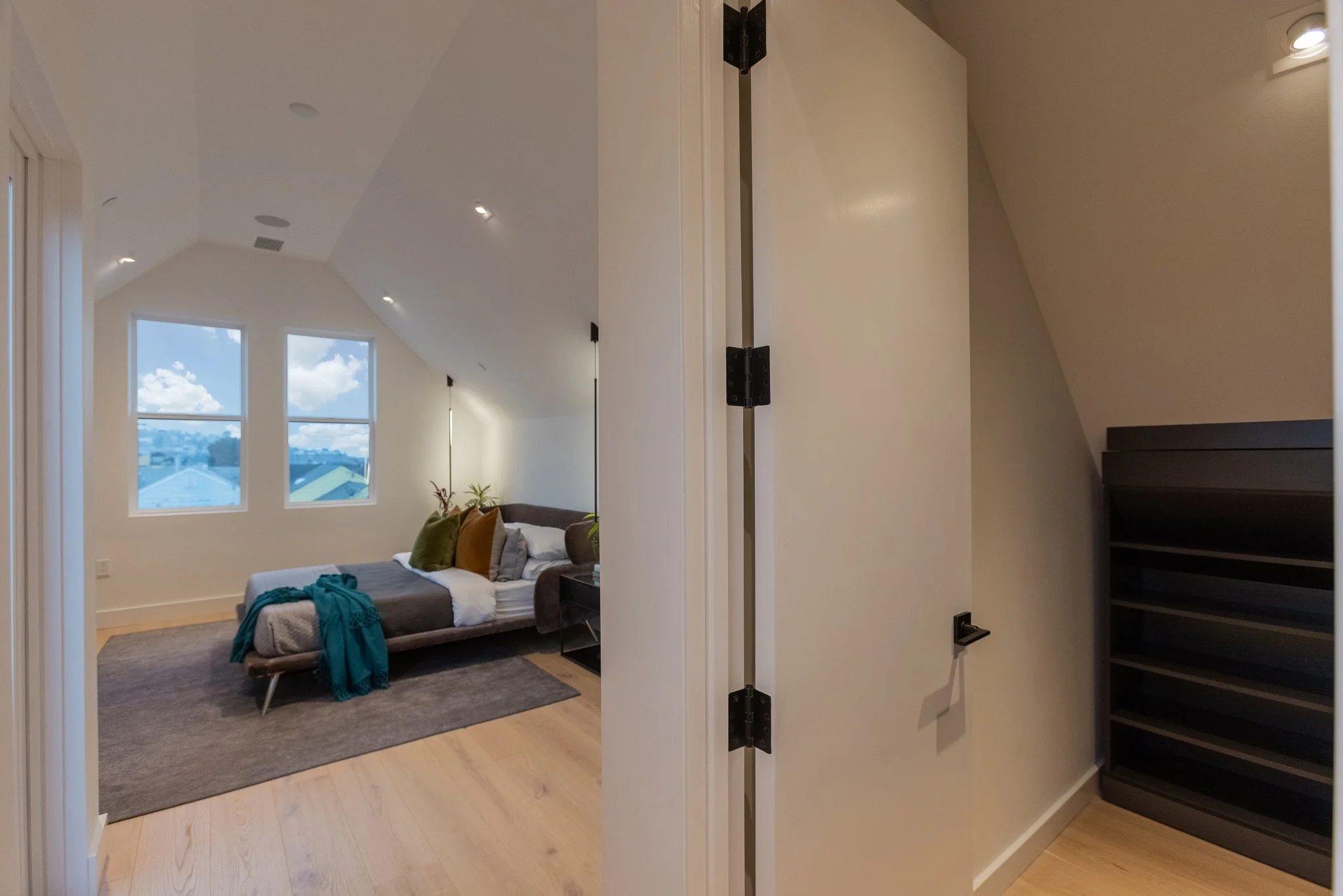 View of a bedroom seen through an open door, with a bed featuring green, yellow, gray, and white pillows, large windows showing a partly cloudy sky, and furniture including a black nightstand and bookshelf.