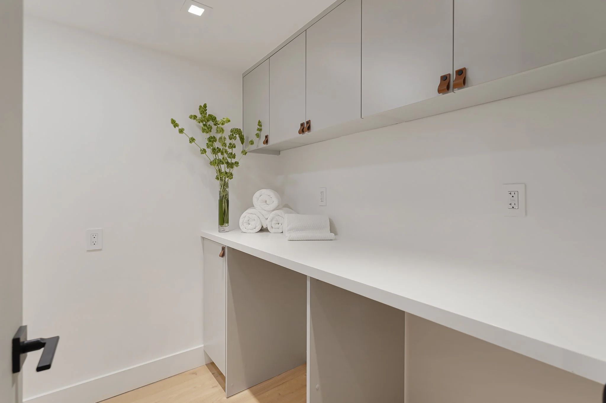 A minimalist laundry or utility room with white cabinetry and a white countertop. On the countertop, there are three rolled white towels and a single folded towel, along with a vase containing green foliage. The room features white walls, light wood 