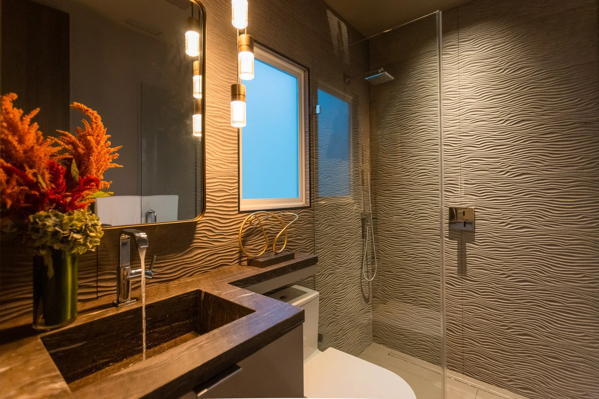 Modern bathroom with textured beige walls, a window, a shower with glass partition, a brown counter with a vessel sink, flowers in a vase, and contemporary lighting fixtures.