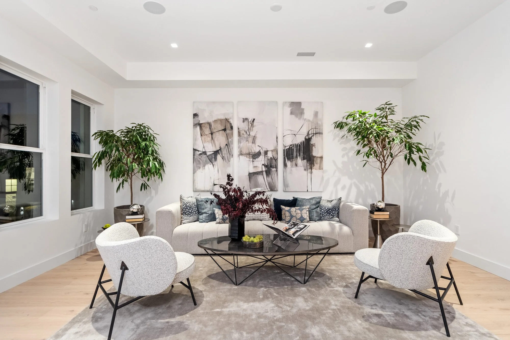 Modern living room with white walls, beige sofa with blue and white pillows, four white chairs, a black coffee table with a flower arrangement, abstract art on the wall, and two tall potted plants.