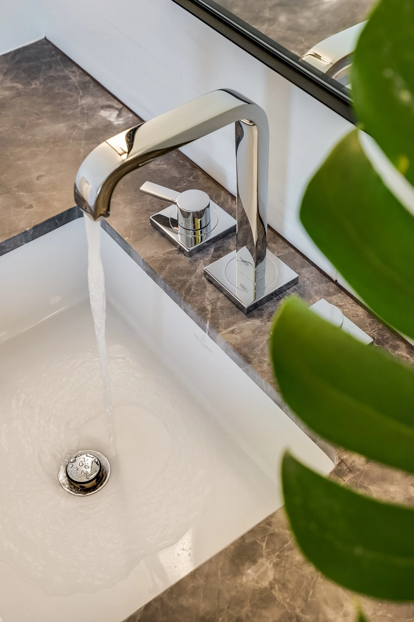 Close-up of a modern kitchen sink with a chrome faucet, water flowing into a white basin, and a nearby green plant.