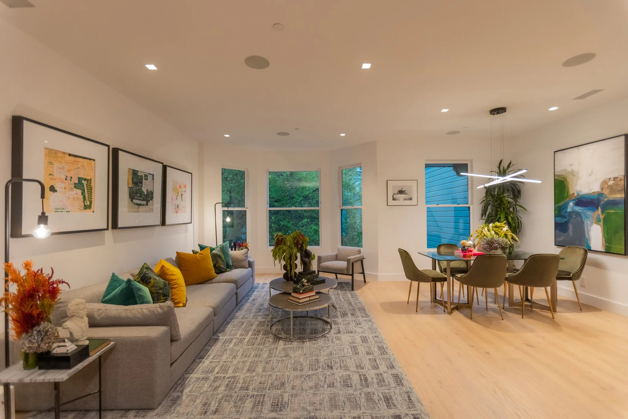 Modern living room and dining area with large window, green plants, colorful artwork, a gray sofa with multicolored pillows, a round coffee table with books and a plant, a dining table with six chairs, and contemporary lighting fixtures.