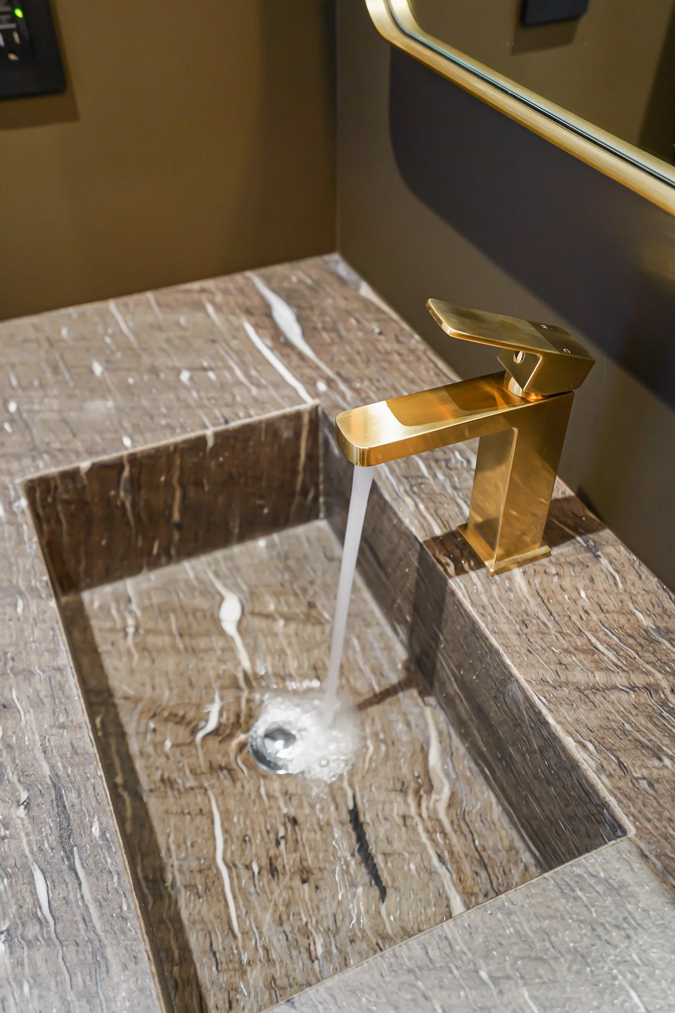 A modern bathroom sink with a gold faucet and running water, set against brown marble countertops and a mirror.
