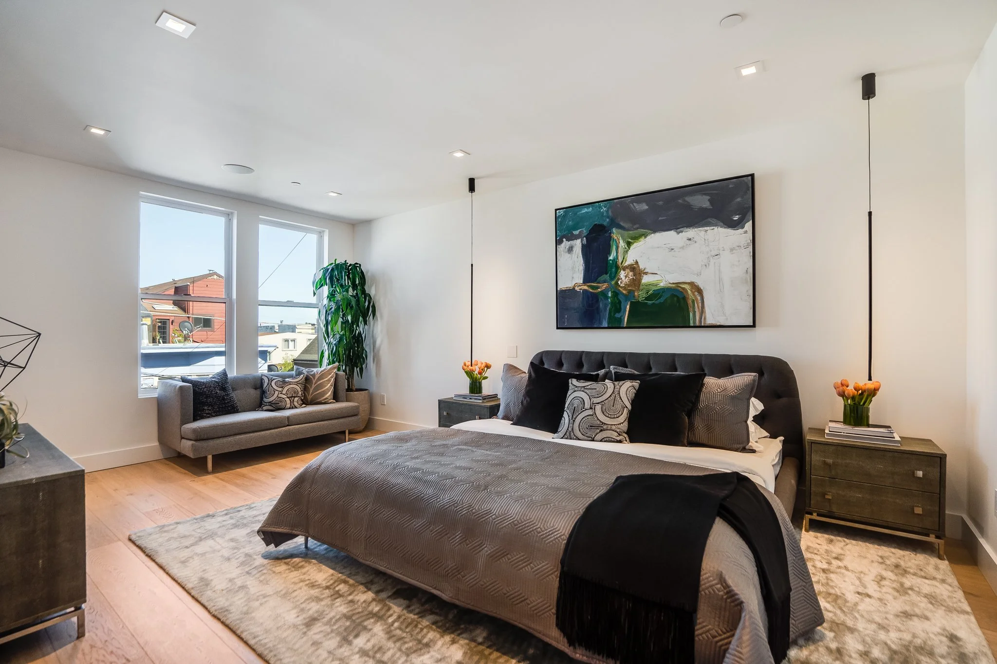 Modern bedroom with a large bed, decorative pillows, two nightstands with flowers, artwork above the bed, and a window with a view of neighboring buildings.