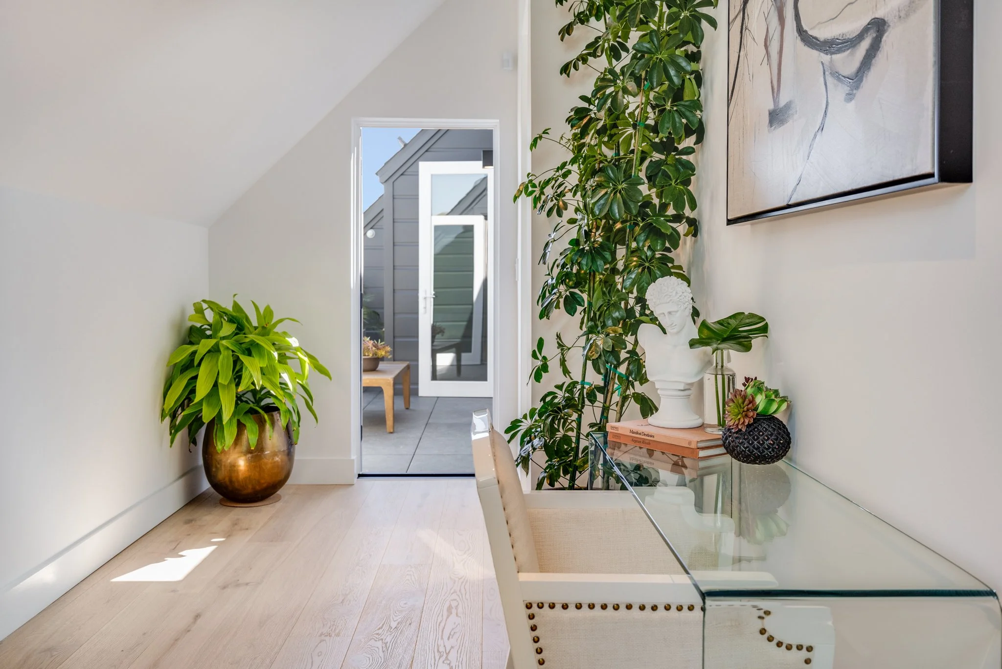 A bright interior space with a glass desk, decorative plants, and art pieces, with a view of a balcony outside.