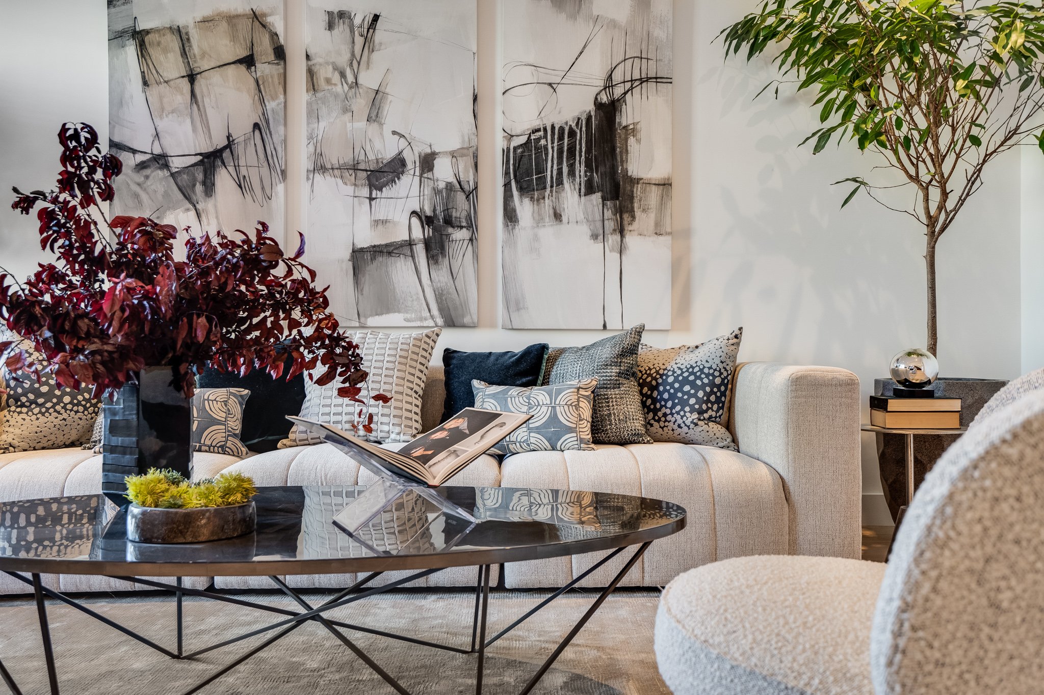 Living room with a beige sofa decorated with patterned cushions, a black coffee table with a book and a vase of dark red leaves, a side table with books and a glass decor object, a large abstract black and white painting on the wall, a potted tree in