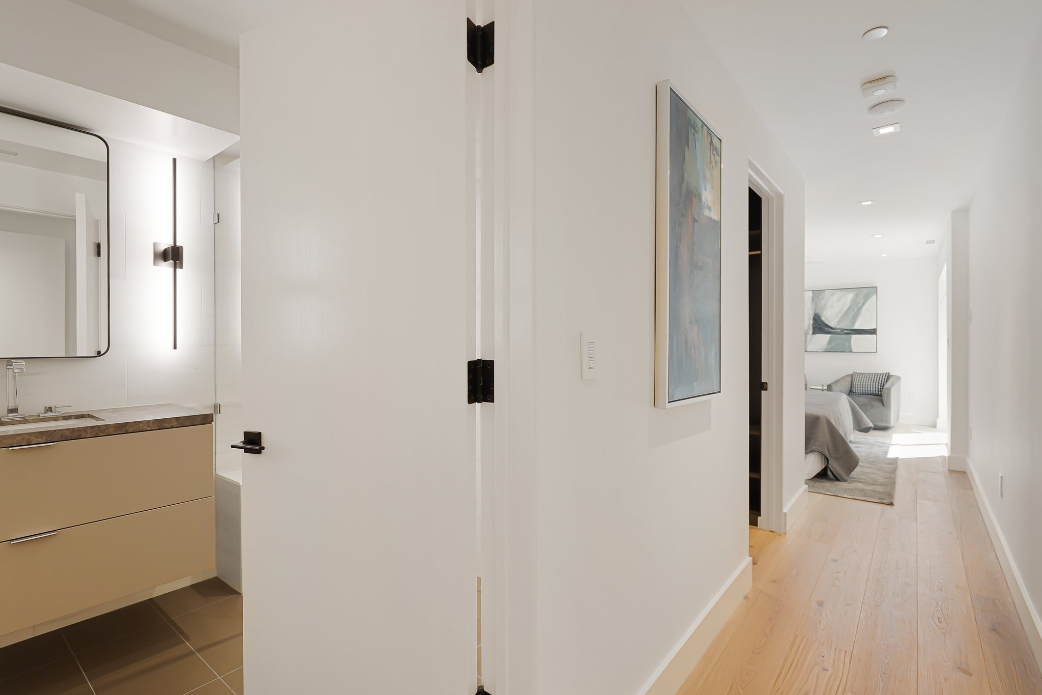 Modern interior view of a hallway with white walls, light wood flooring, and artwork, leading to a living area with a sofa and large window.