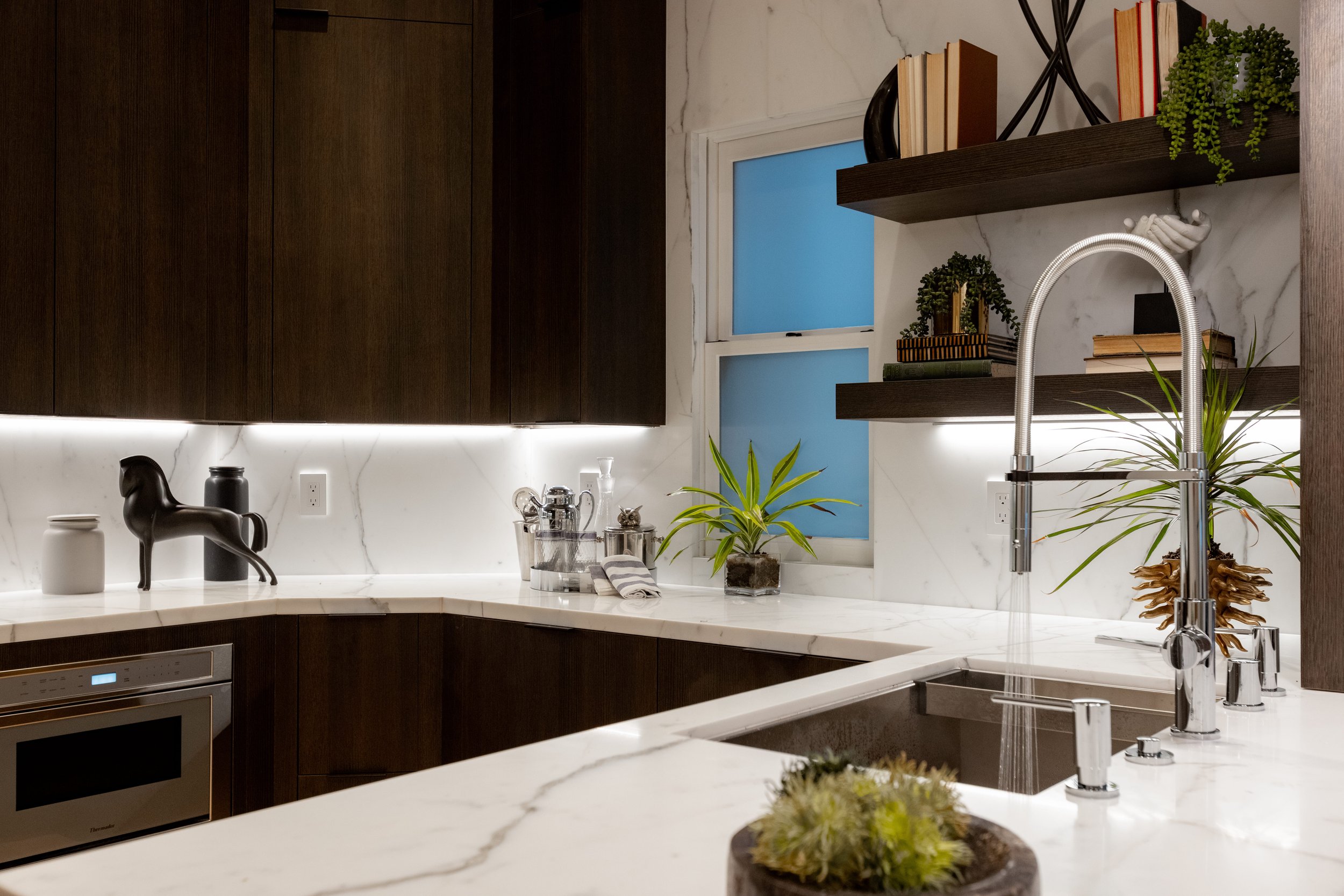 Modern kitchen with white marble countertops, dark wood cabinets, and open shelves with books and plants. A window has a blue tint, and a stainless steel faucet is over a sink. Decorative items and potted plants are on the counter.