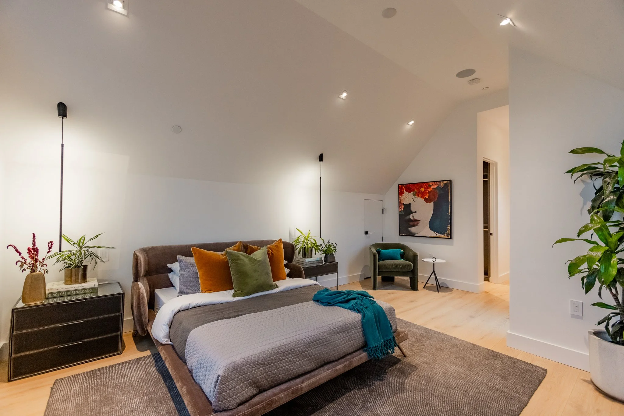 Modern bedroom with a bed featuring multiple colorful pillows, a blanket, and a throw on top. There are plants on nightstands on either side of the bed. A large abstract portrait hangs on the wall. A small green armchair and a side table are visible 