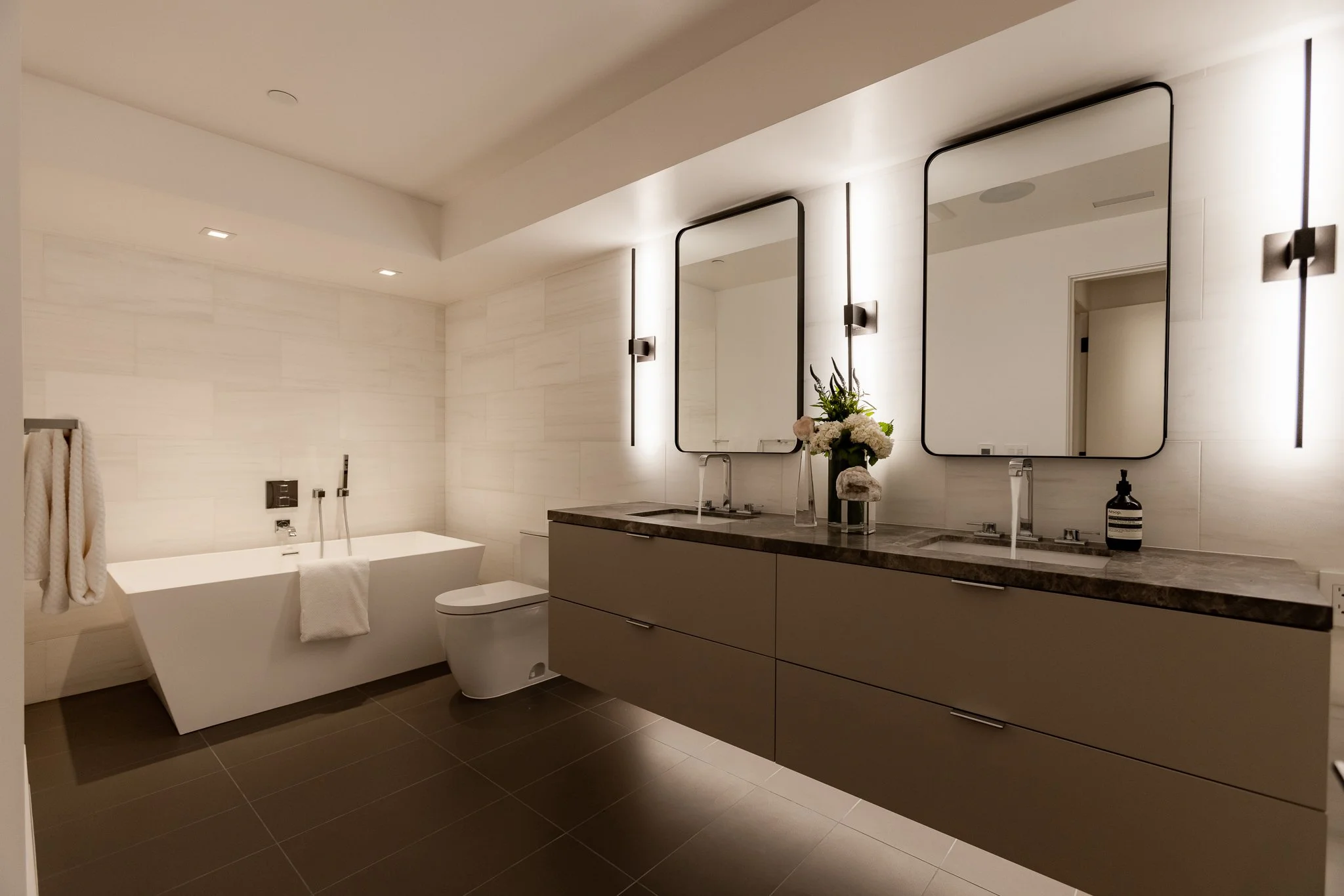 Modern bathroom with white bathtub, double vanity with large mirrors, flowers, and neutral decor.