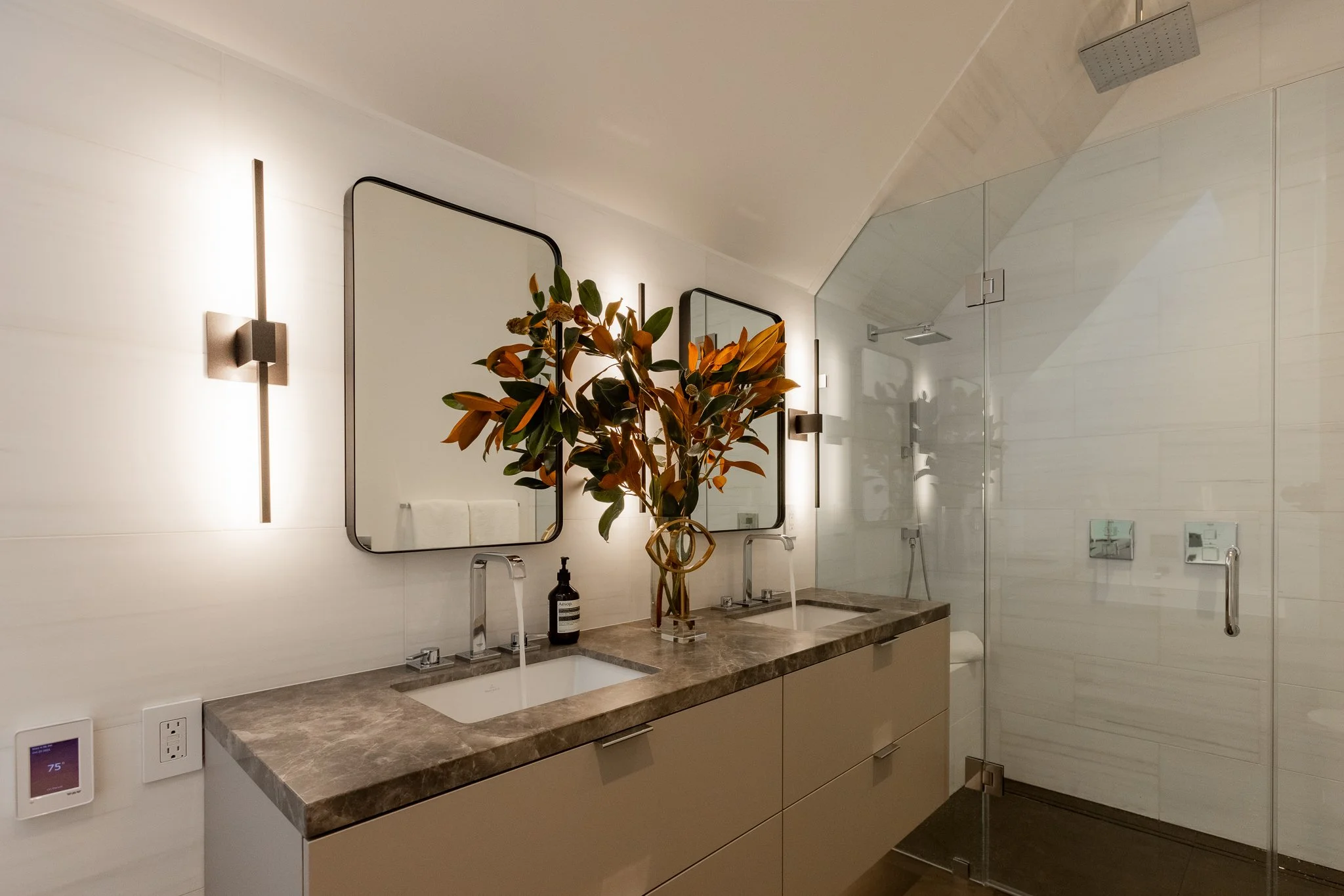 Modern bathroom with double sinks, two mirrors, decorative plant, and a glass-enclosed shower area.