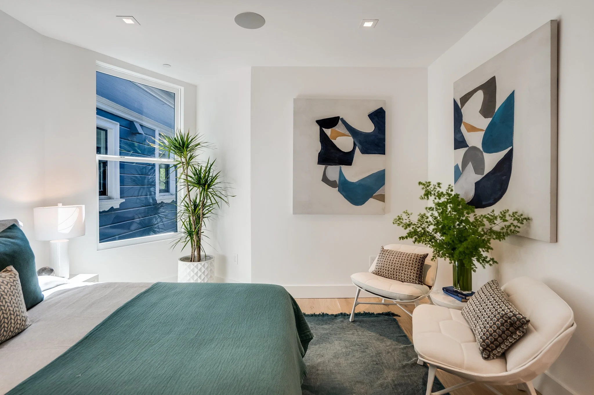 Modern bedroom with white walls, a large window, a bed with green bedding, two armchairs with pillows, abstract artwork on the walls, a potted plant, and a nightstand with a lamp.