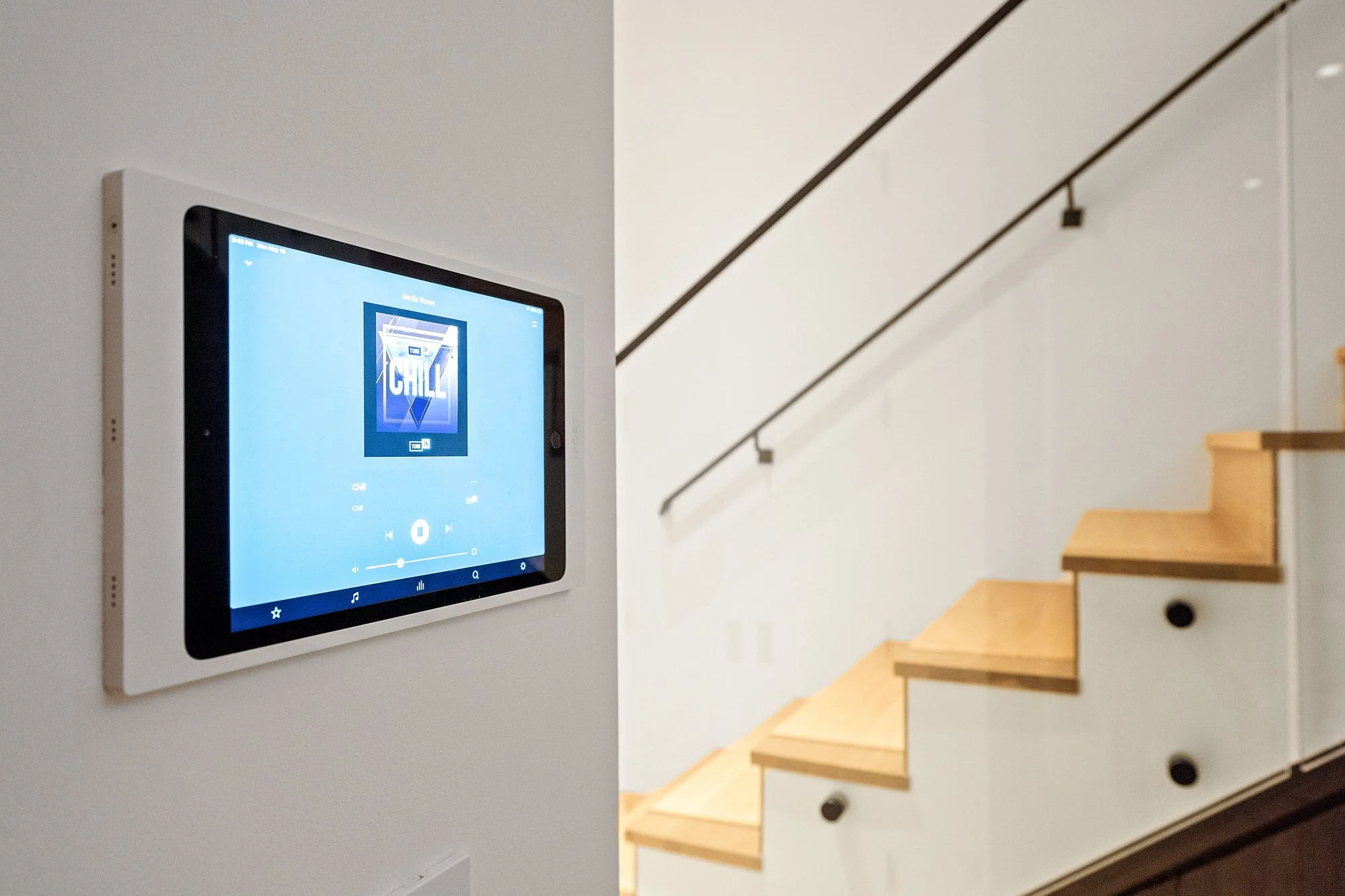 Wall-mounted digital music player displaying a blue screen with the album artwork for 'Chill' and music controls, next to a wooden staircase with black railing.