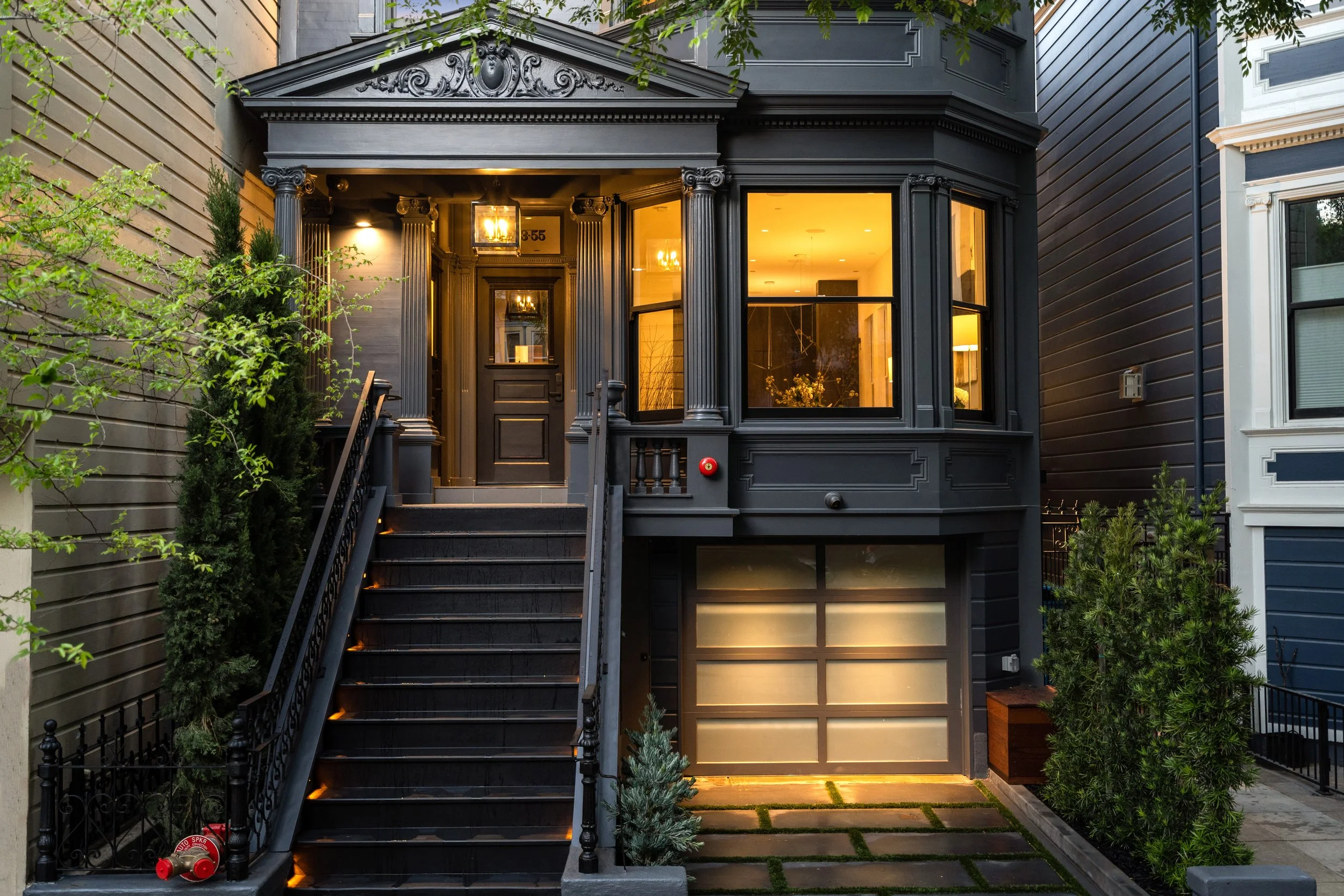 Black Victorian-style house with stairs leading to a front door, large bay windows, and lush green trees and shrubs in the front yard.