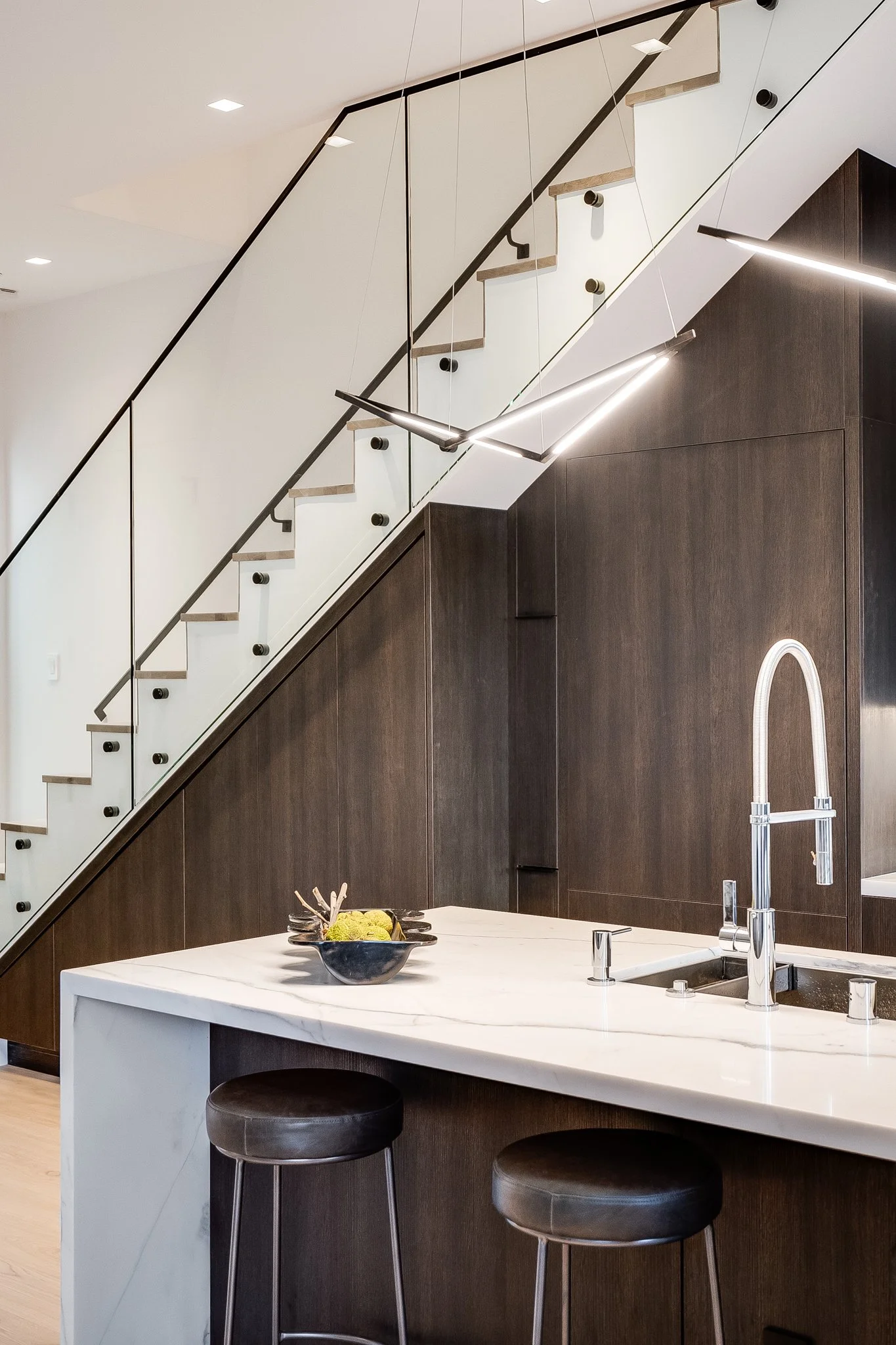 Modern kitchen with a white marble island, dark wood cabinets, and a staircase with glass and metal railing.