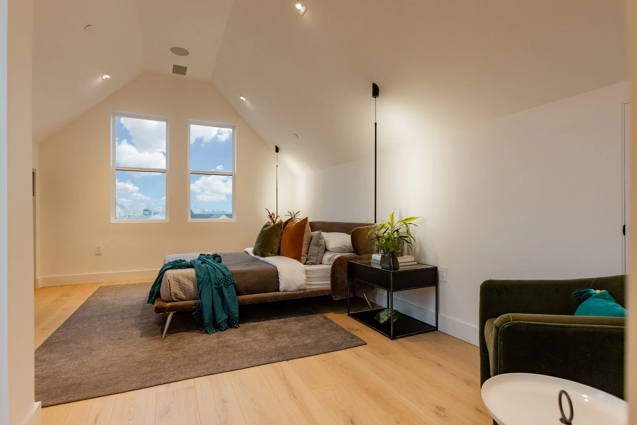 A modern bedroom with a sloped ceiling, featuring a bed with colorful pillows, a black nightstand with a plant, window view of a blue sky with clouds, and a green armchair.