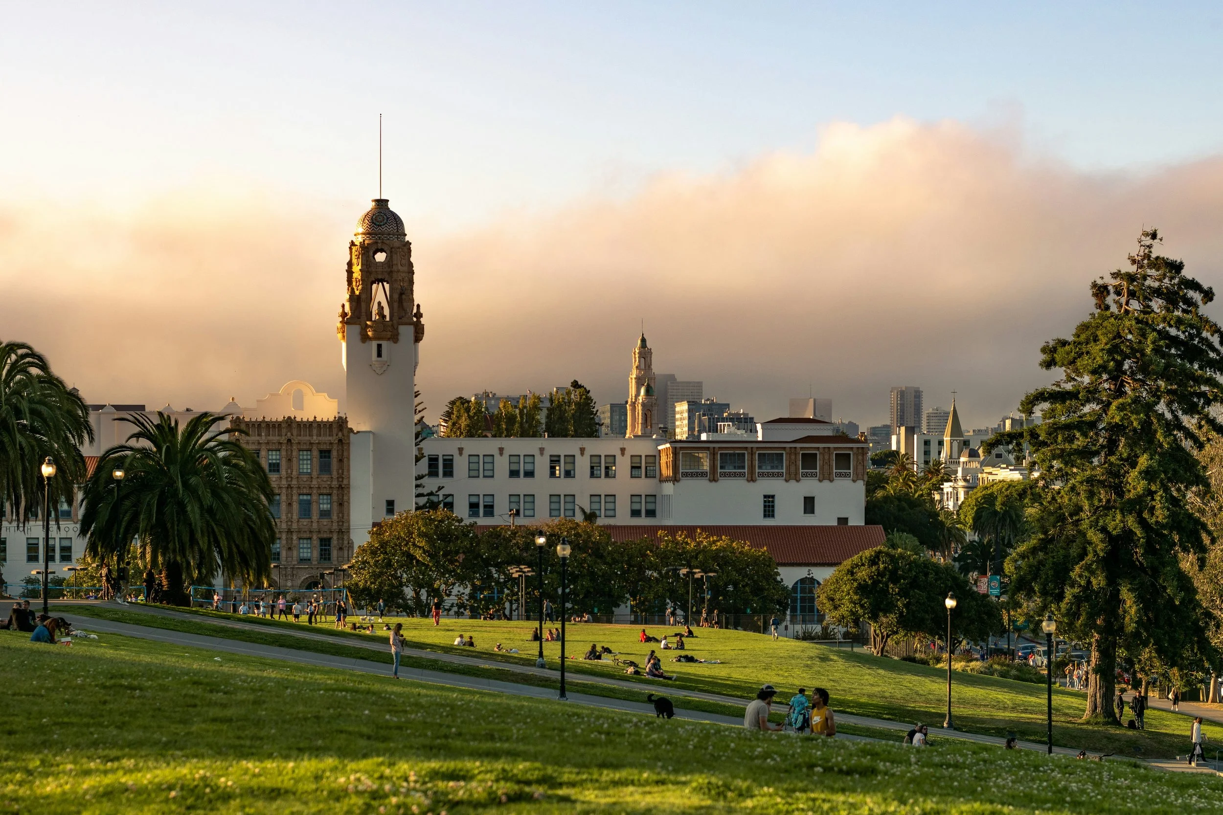 A city park with people sitting and walking on a grassy area, trees, and a historic white building with a clock tower, with a skyline of modern buildings in the background.