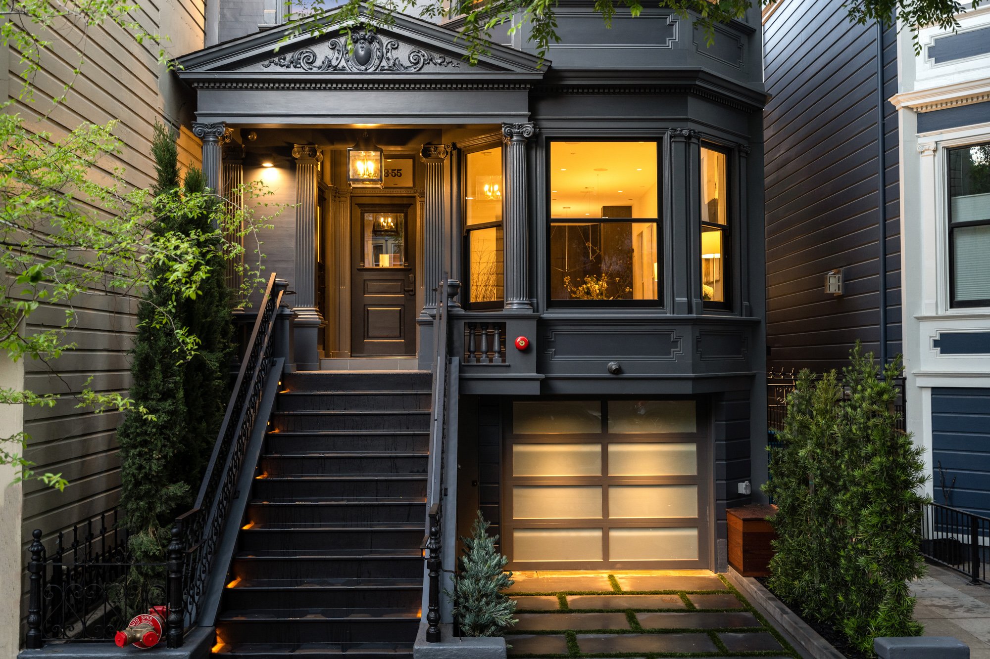 A black Victorian-style house with a bay window and a front porch accessed by stairs. The house has interior lighting visible through large windows and exterior lighting on the stairs and porch. There are trees and shrubs on either side of the stairs