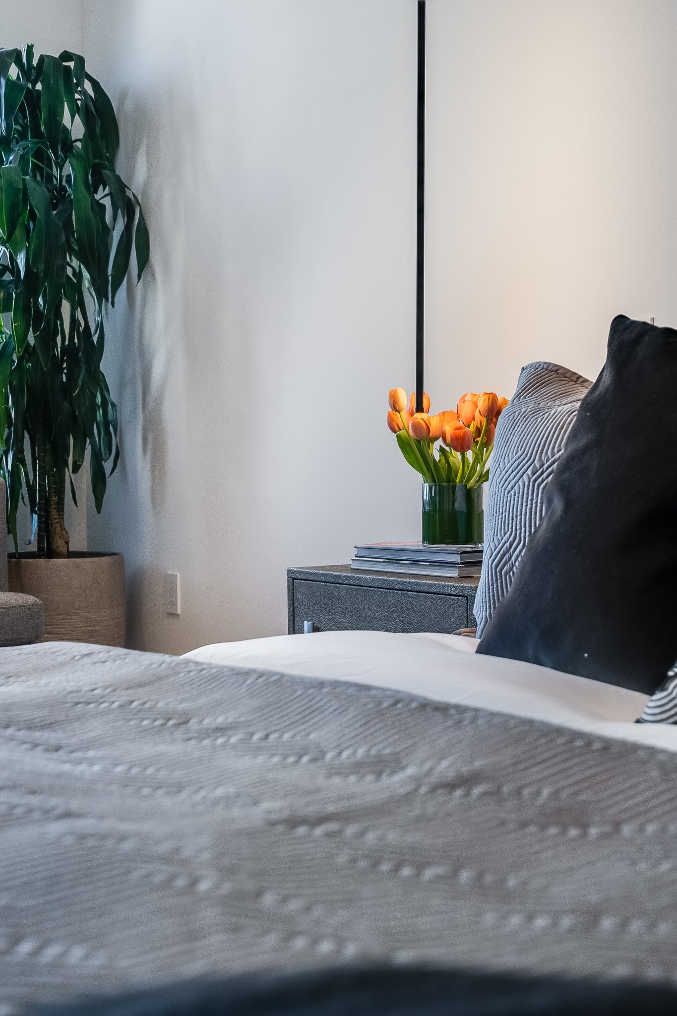 A bedroom with a bed that has gray and black pillows, a nightstand with a stack of books and a vase of orange tulips, a tall green houseplant in a beige pot, and a wall-mounted black lamp.