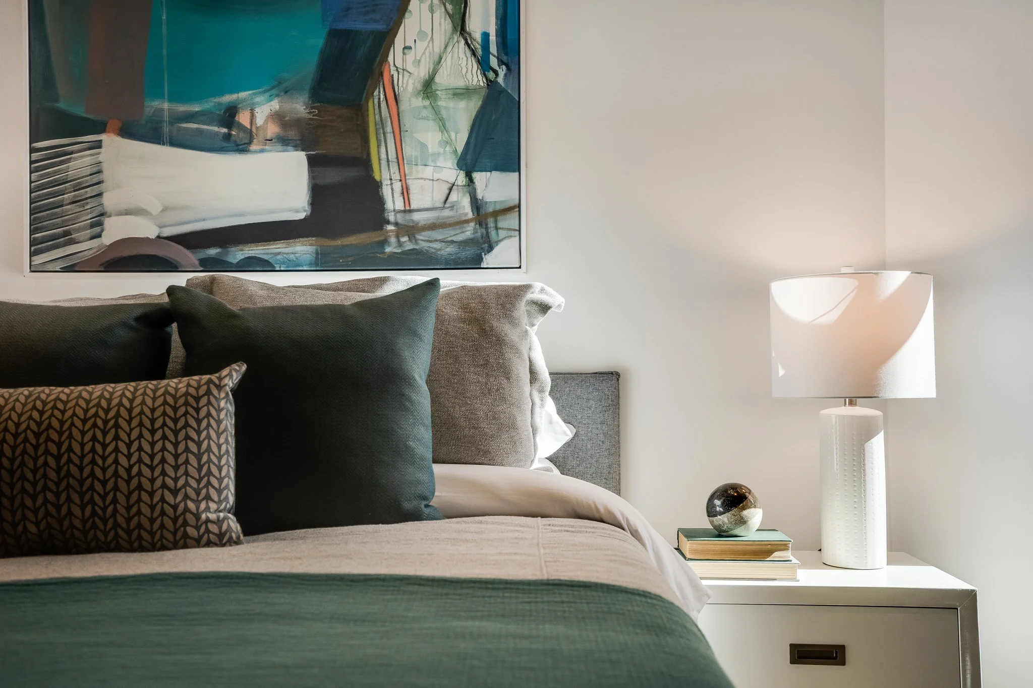 A neatly made bed with multiple pillows in shades of dark green, beige, and patterned in black and beige. There is a white bedside table with a white textured lamp, a globe, and a few stacked books. Above the bed hangs a large, abstract painting with