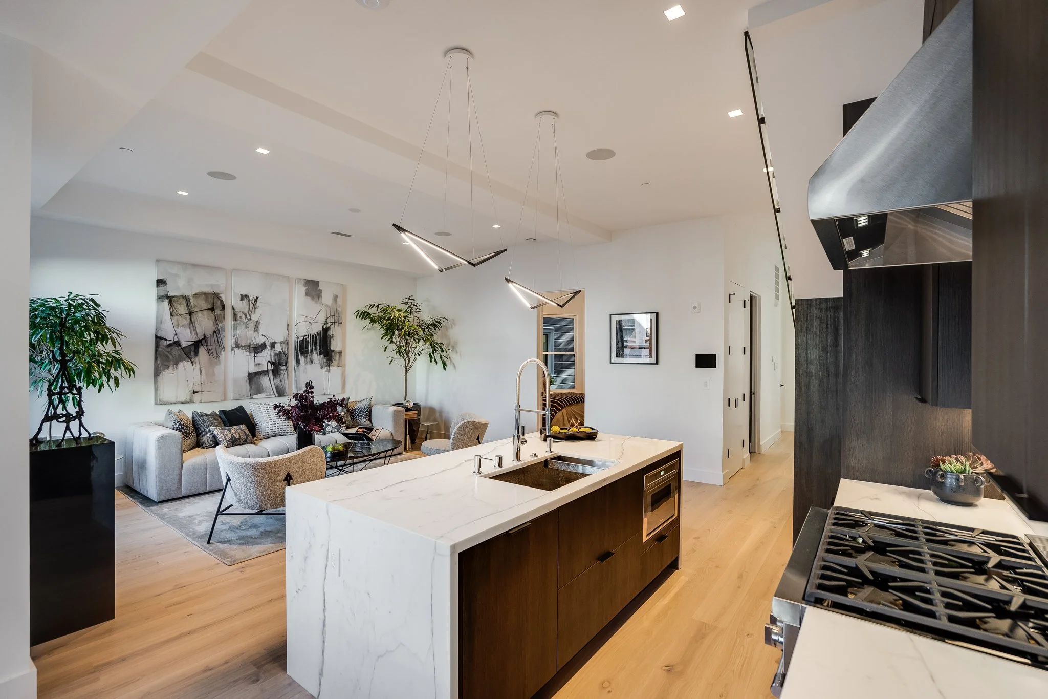 Open-concept living room and kitchen with a white marble island, black cabinetry, and modern lighting fixtures. The living area has a white sofa, patterned pillows, two armchairs, abstract wall art, and potted plants.