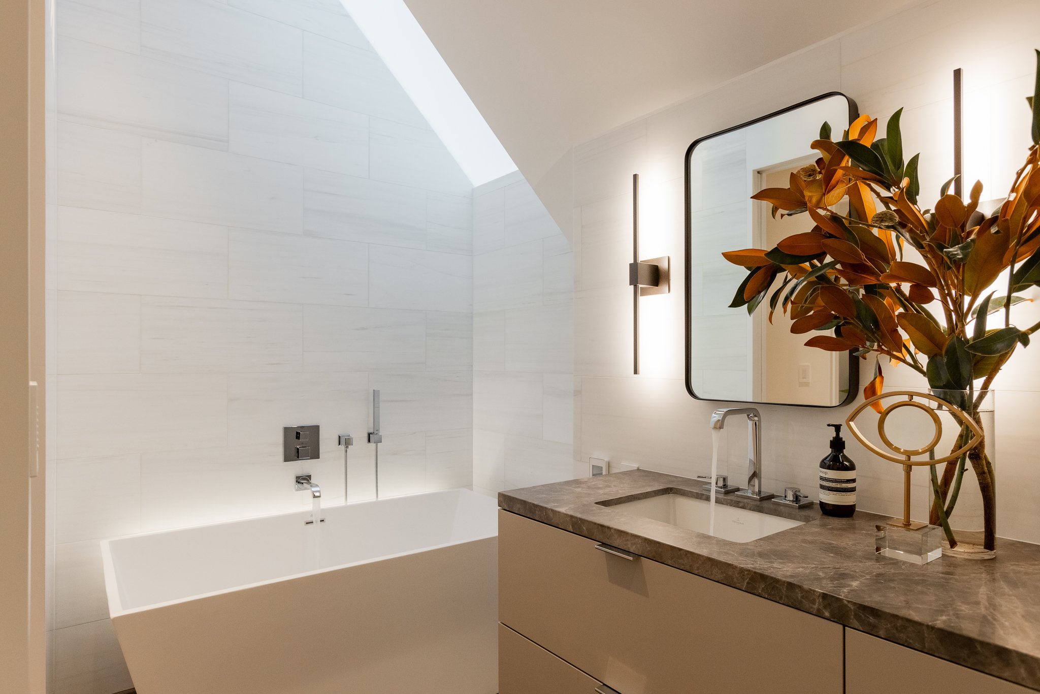 Modern bathroom with a white bathtub, a marble vanity with a sink, a large mirror, a wall sconce, a glass vase with lush foliage, and a soap dispenser, all illuminated by natural light from a skylight.