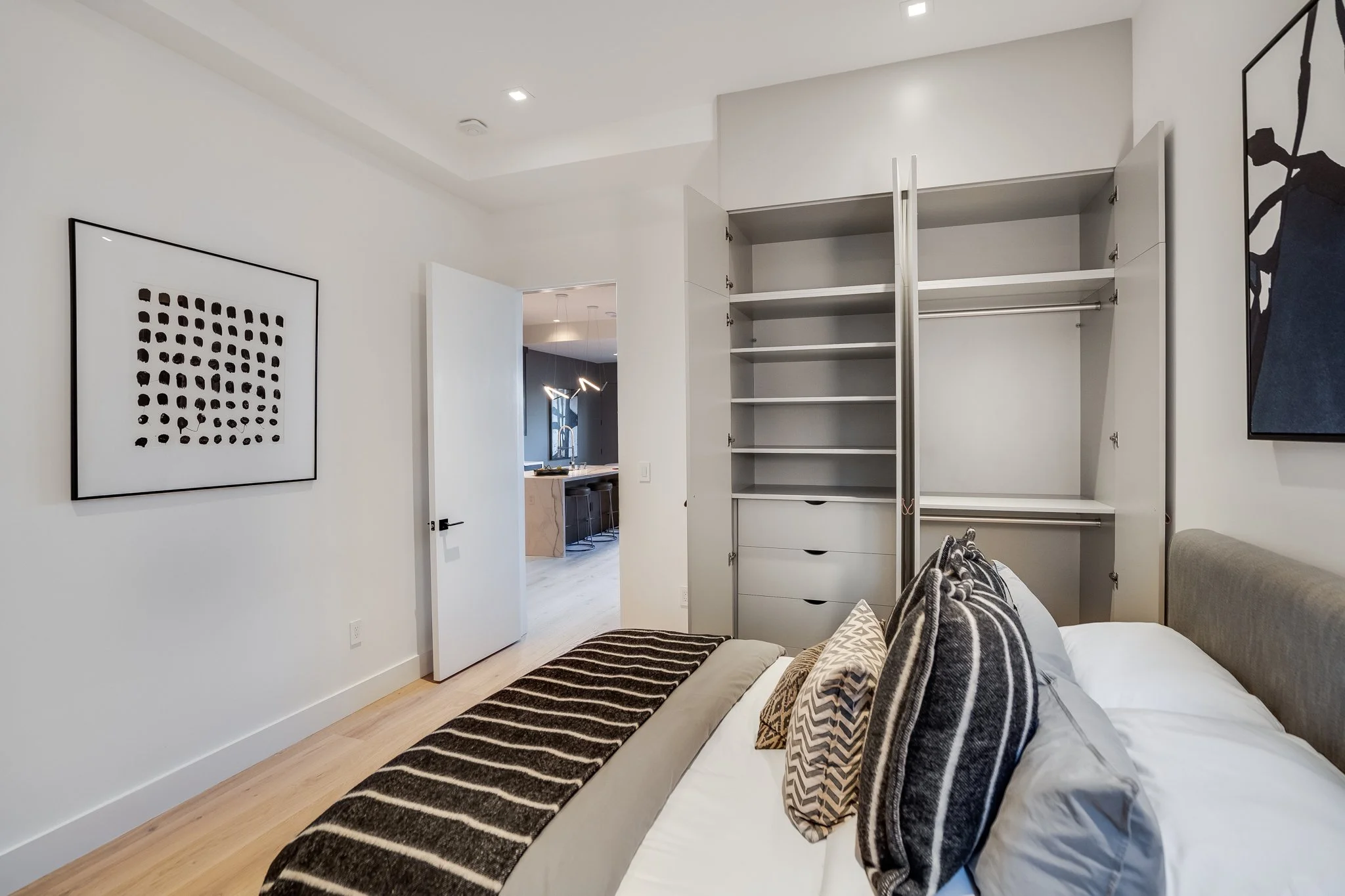 Bedroom with bed, pillows, open closet, artwork on walls, door open to living area and kitchen.
