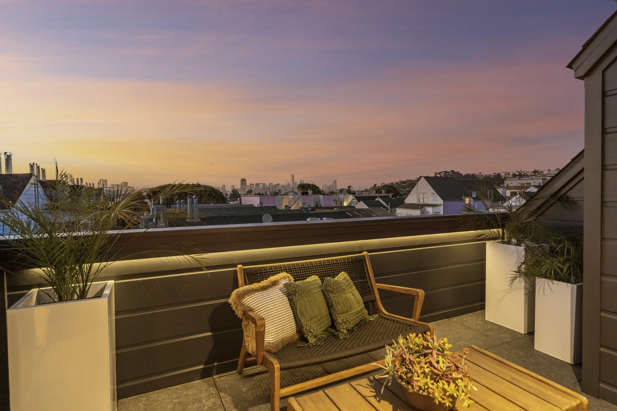 Outdoor balcony with a wooden bench and green cushions, potted plants, and a sunset sky with pink and purple hues.