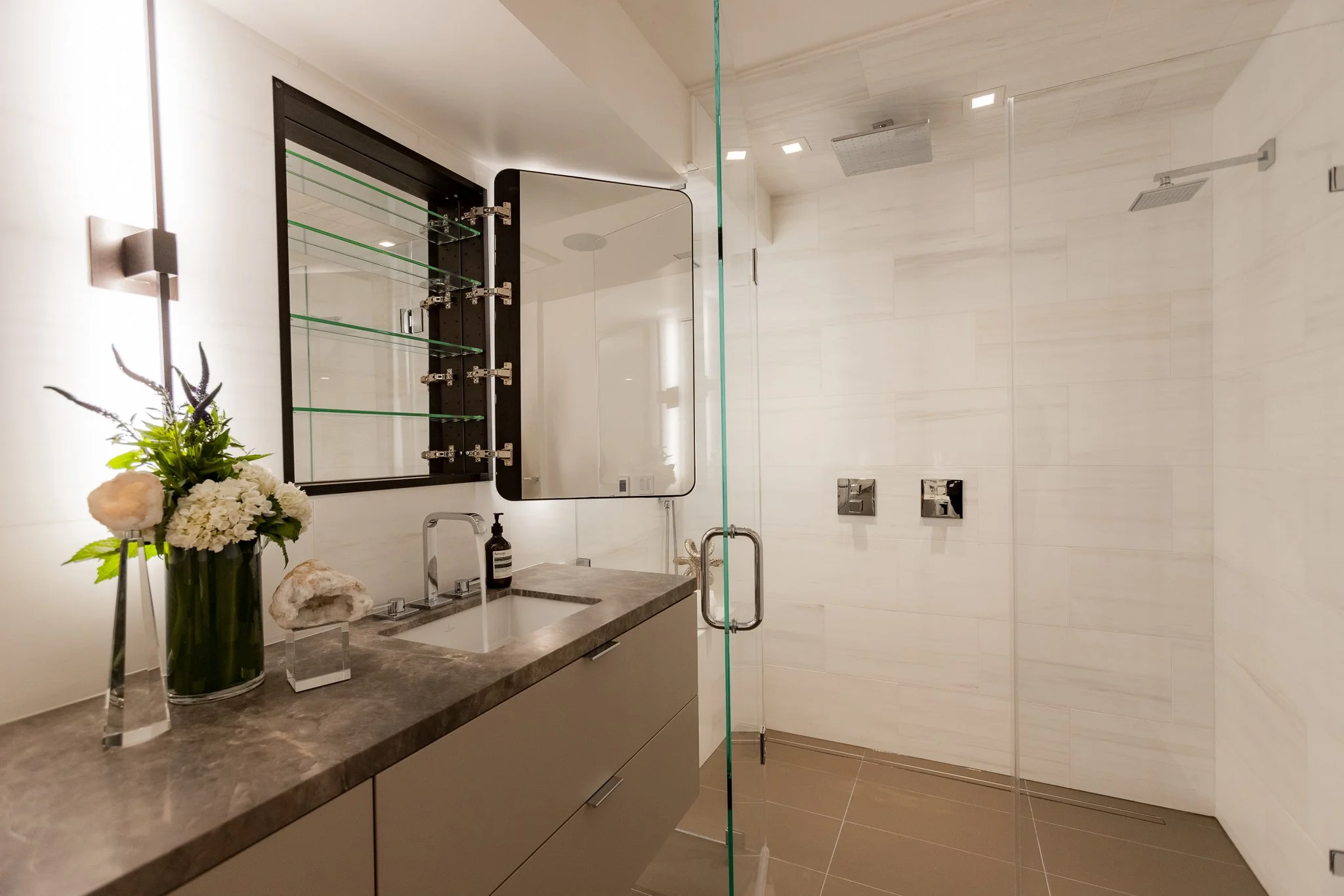 Modern bathroom with a beige countertop, flowers, a soap dispenser, mirror, glass shower enclosure, two shower controls, a large overhead shower, and white tiled walls.