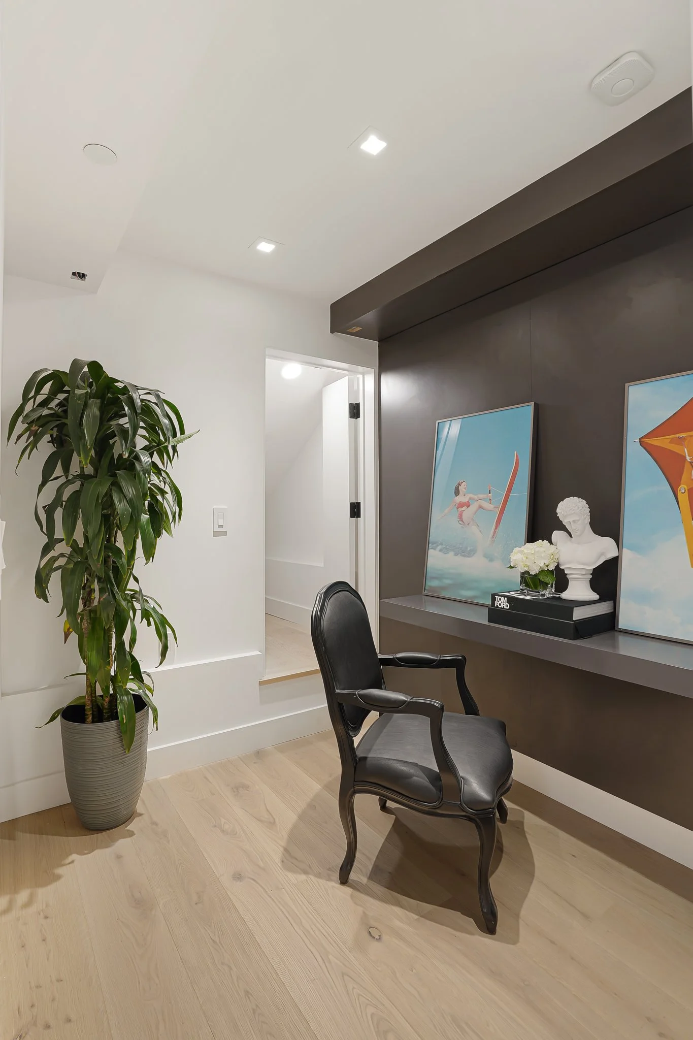 A modern home office corner with a tall potted plant, a black leather armchair, and a dark wall shelf with ocean-themed artwork, a white bust sculpture, and a flower arrangement.