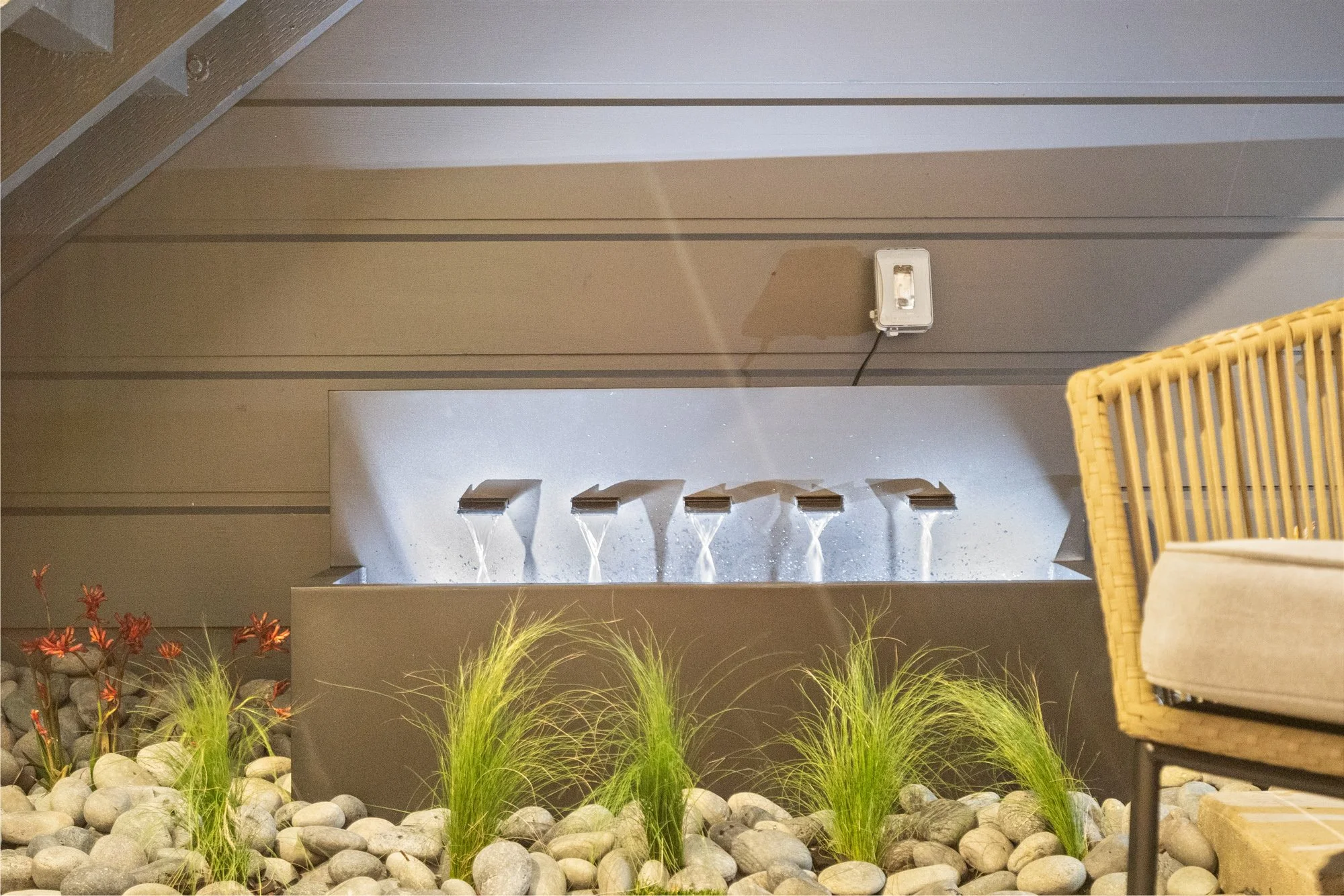Indoor water feature with four spouts flowing water into a rectangular container, decorated with rocks and tall grasses, next to a wicker chair with a cushion.