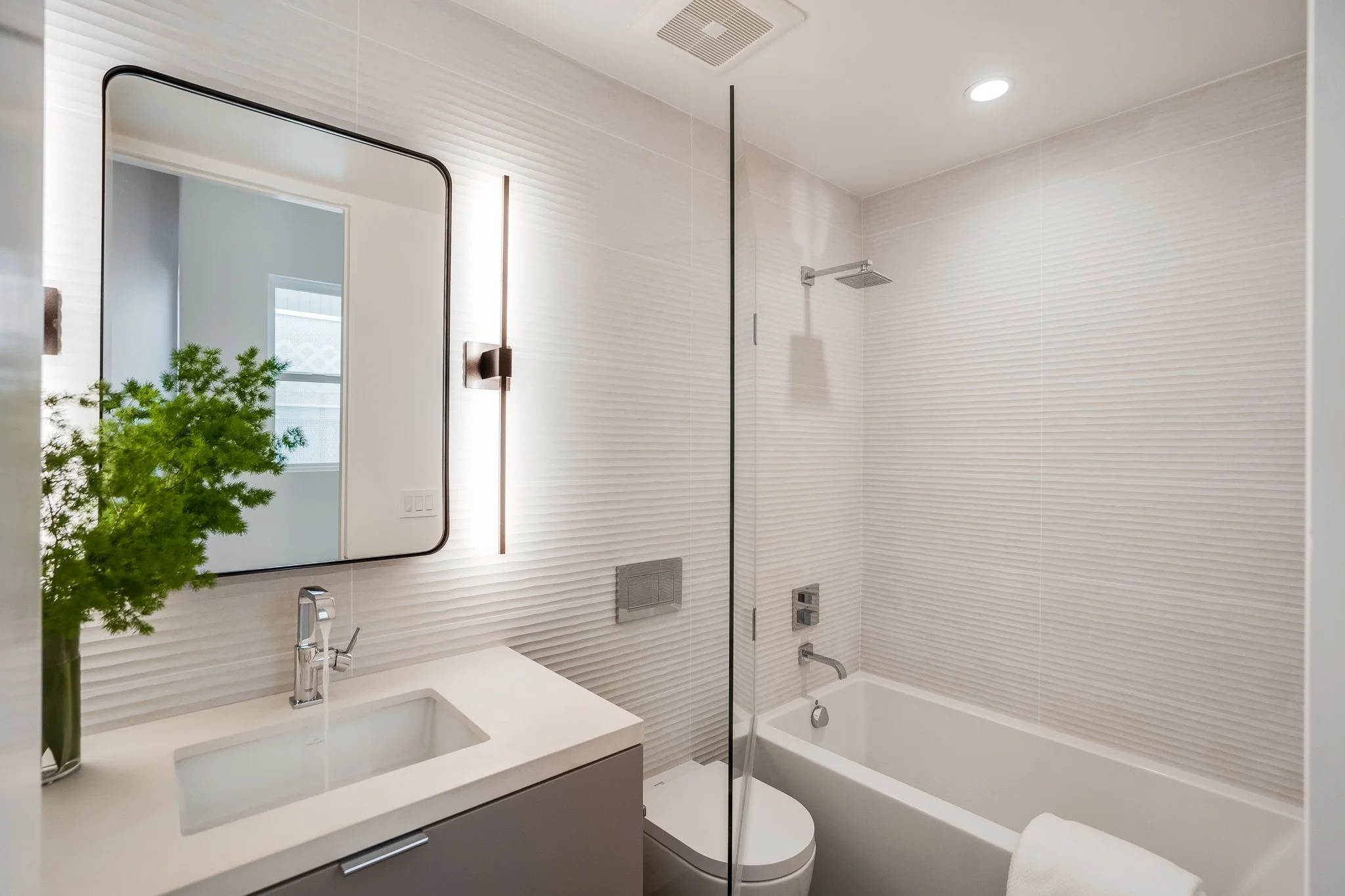 Modern bathroom with white textured walls, a large rectangular mirror, a white sink with a chrome faucet, a small green plant, a gray vanity cabinet, a toilet, a bathtub, and a glass shower partition.